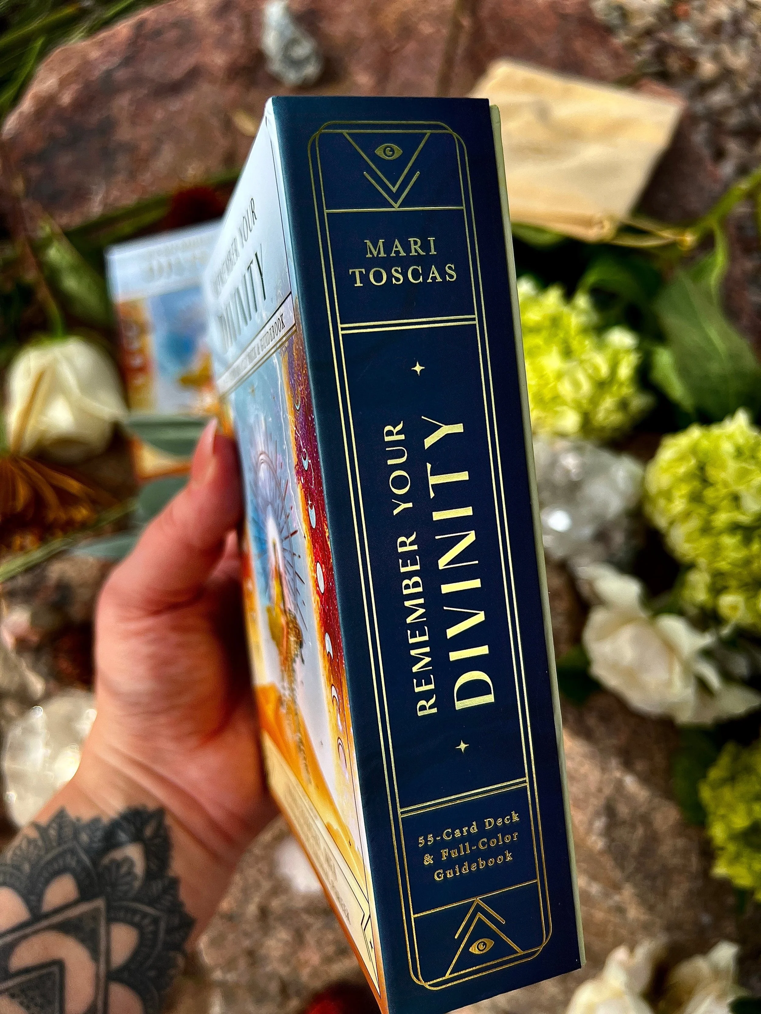 A person holding a deck of 'Remember Your Divinity' tarot cards with a dark blue box and gold lettering, surrounded by flowers and greenery.