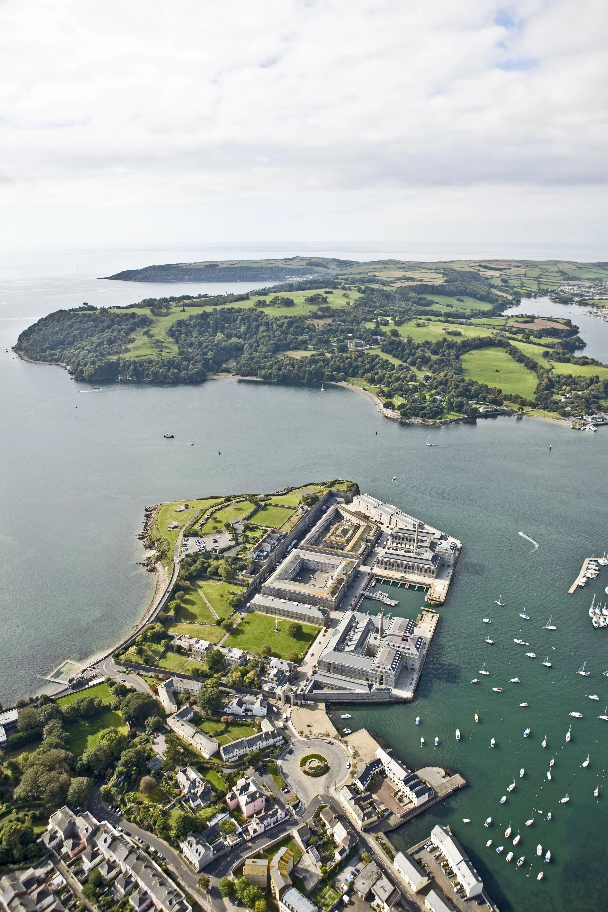 Aerial view of a Plymouth waterfront complex with surrounding green fields, residential buildings, and a marina with boats, near a curved shoreline and distant islands.