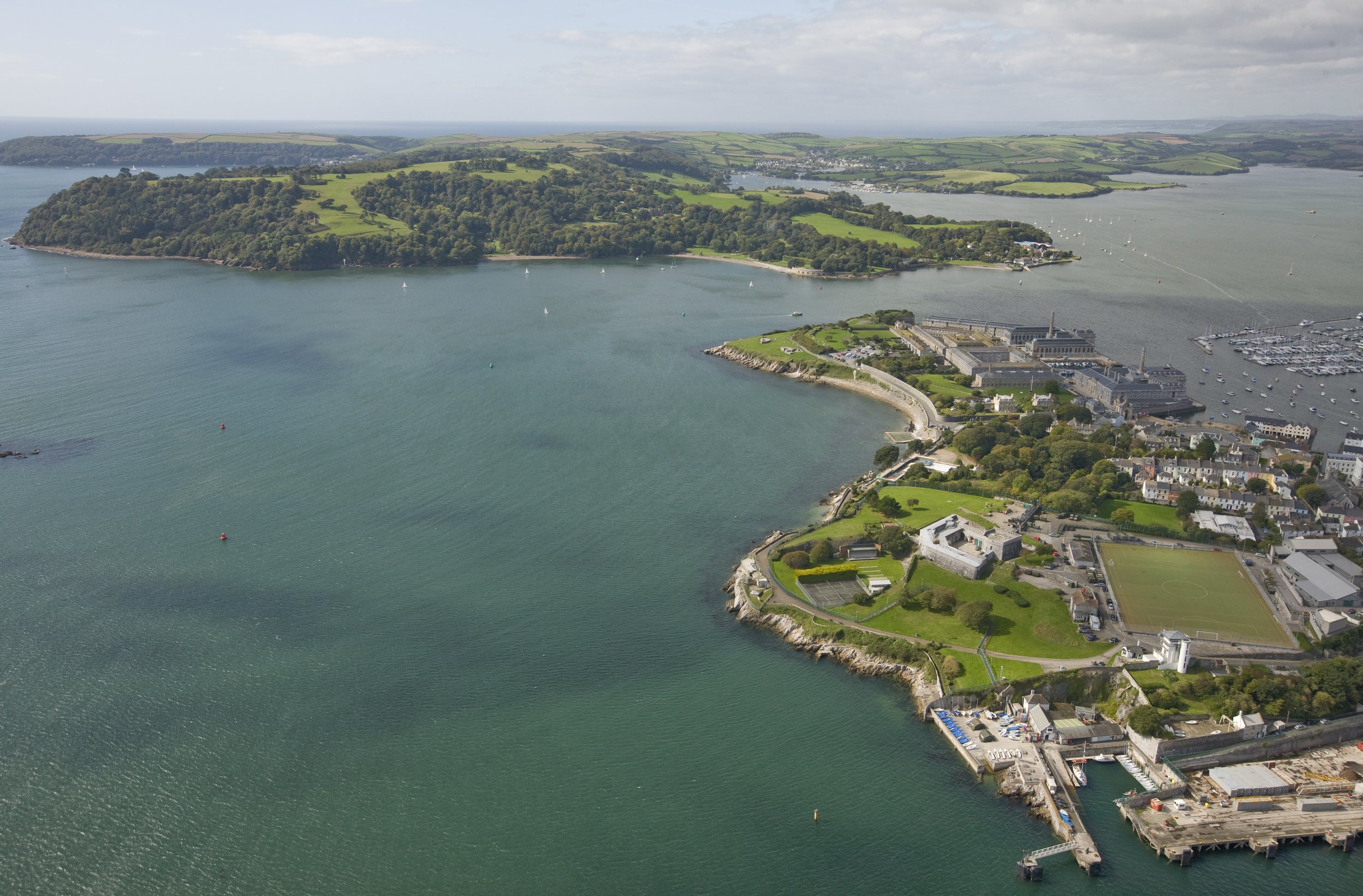 Aerial view of Plymouth, the National Centre for Marine Autonomy, with a harbor, marina, green sports field, historic buildings, and surrounding water with sailboats, lush green hills, and distant land masses.