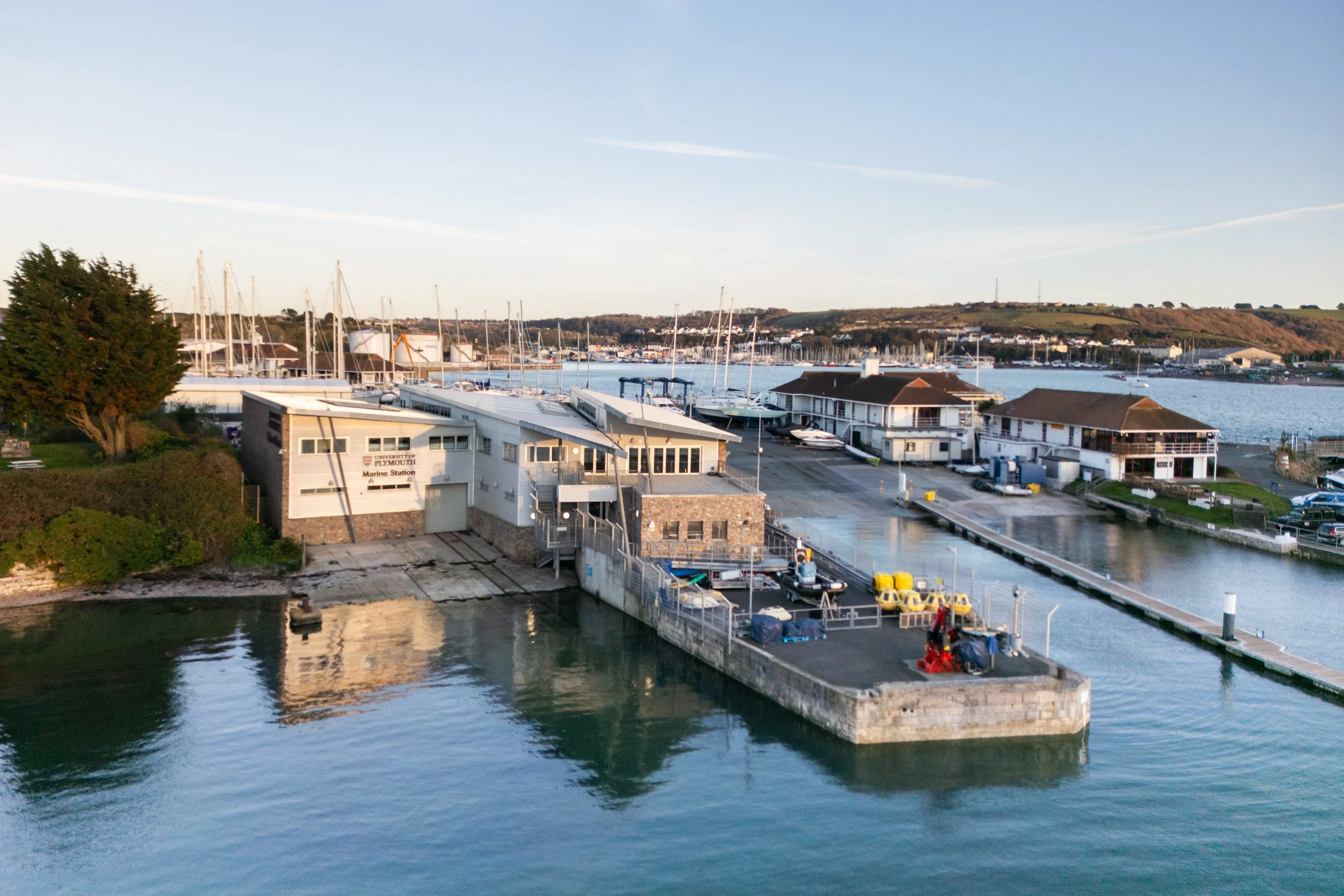 Aerial view of University of Plymouth Marine Station with boats docked, buildings, and a calm body of water reflecing the structures during daytime.