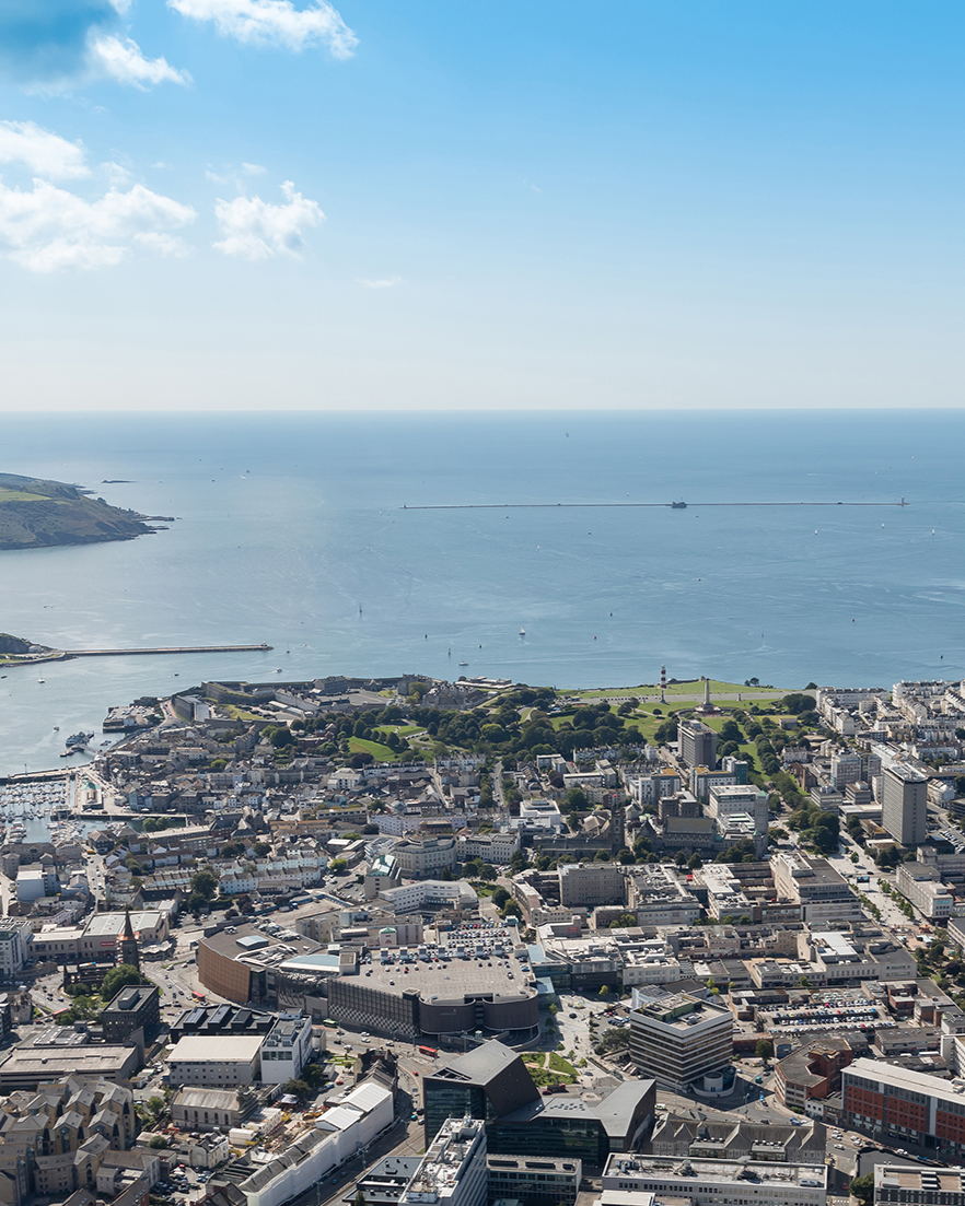 Aerial view of Plymouth with buildings, roads, and green parks, overlooking the ocean with a clear blue sky.