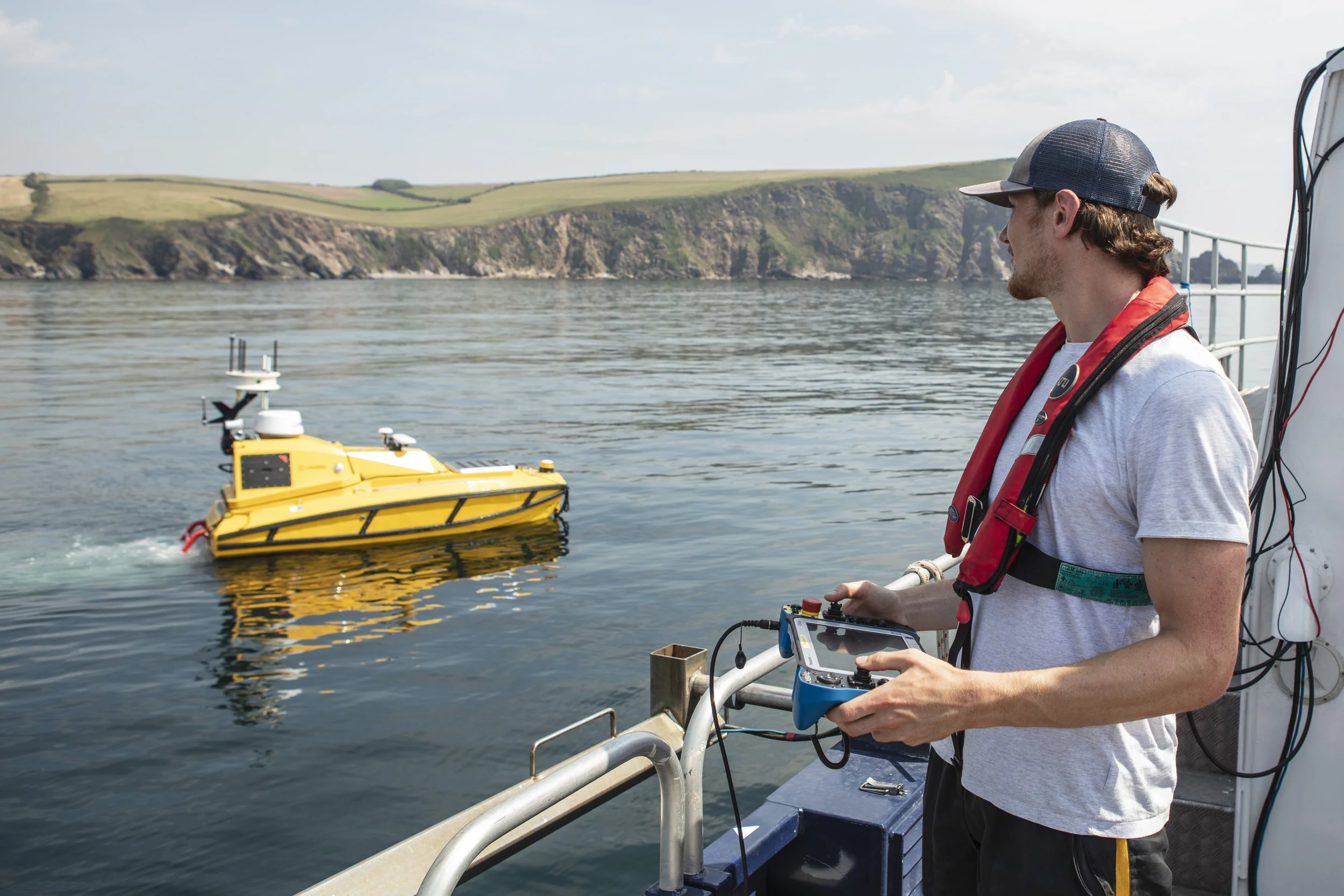 A young man on a boat controls CETUS- a yellow autonomous surface vessel with a tablet, on a body of water with cliffs and greenery in the background.