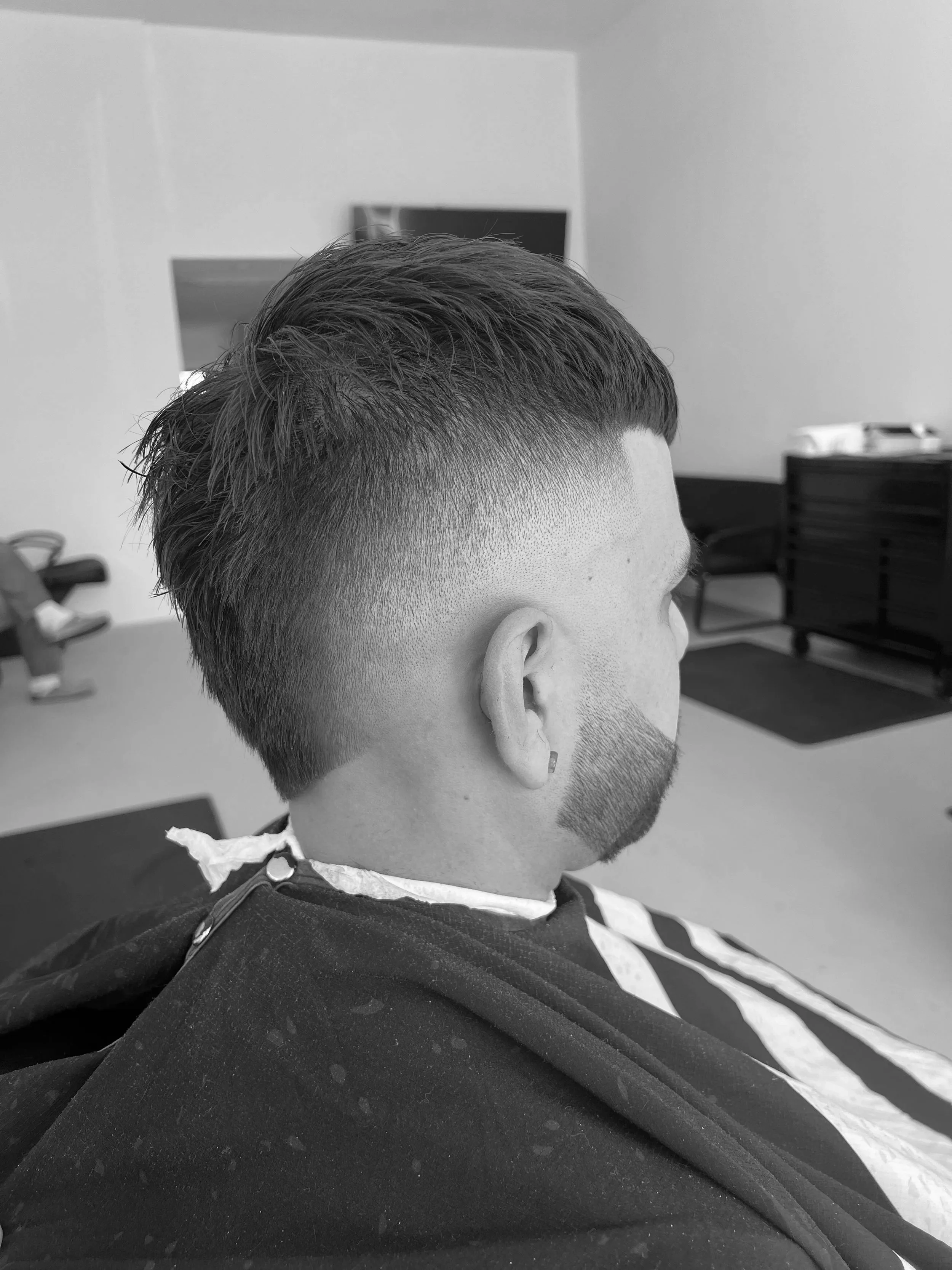 A man with a fresh haircut sitting in a barbershop chair, with style fading on the sides and longer hair on top, in a black-and-white photo.