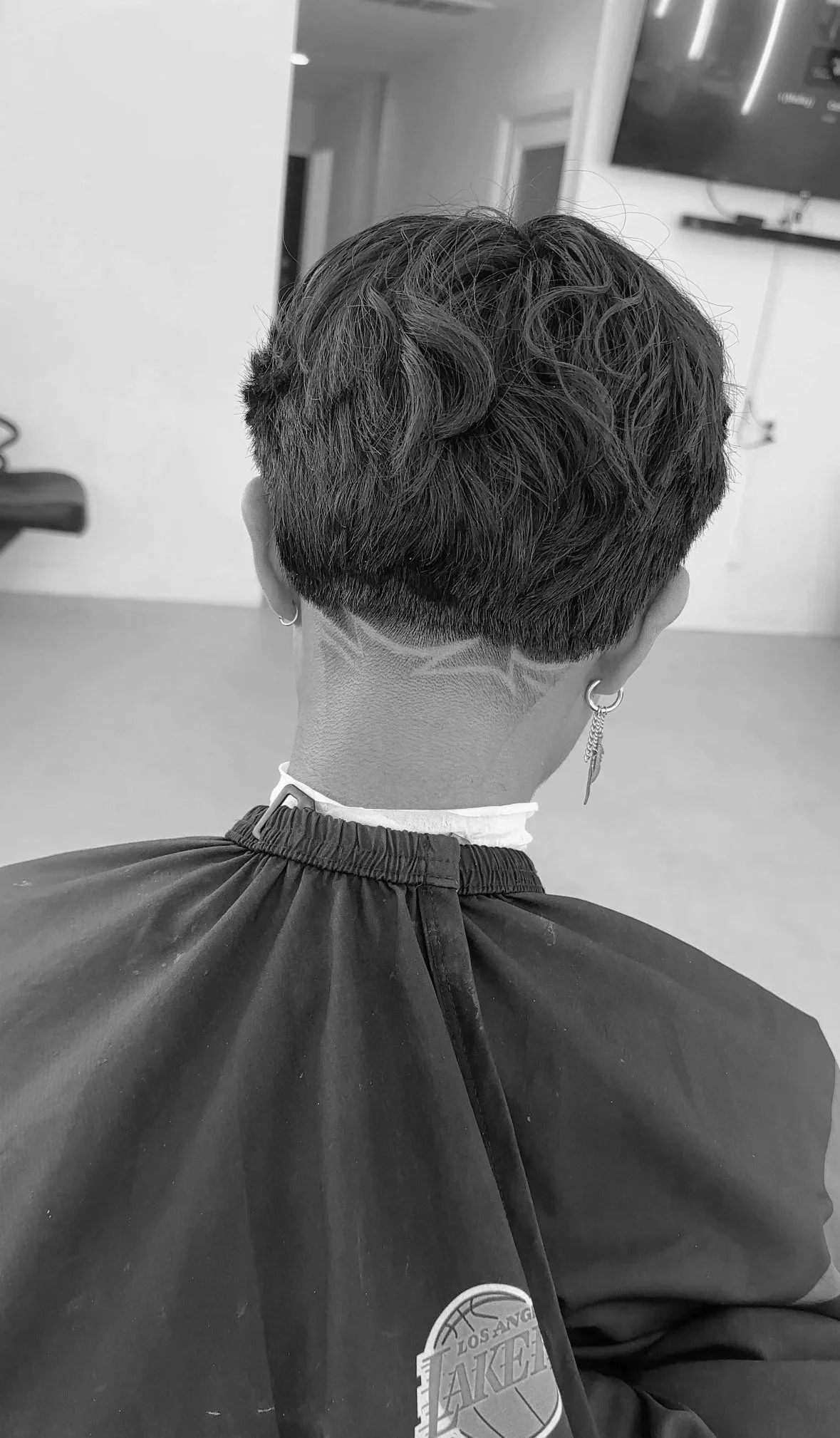 Back of a person with short, curly hair with a shaved design, wearing earrings and a Lakers jacket, sitting in a salon chair in a hair salon.