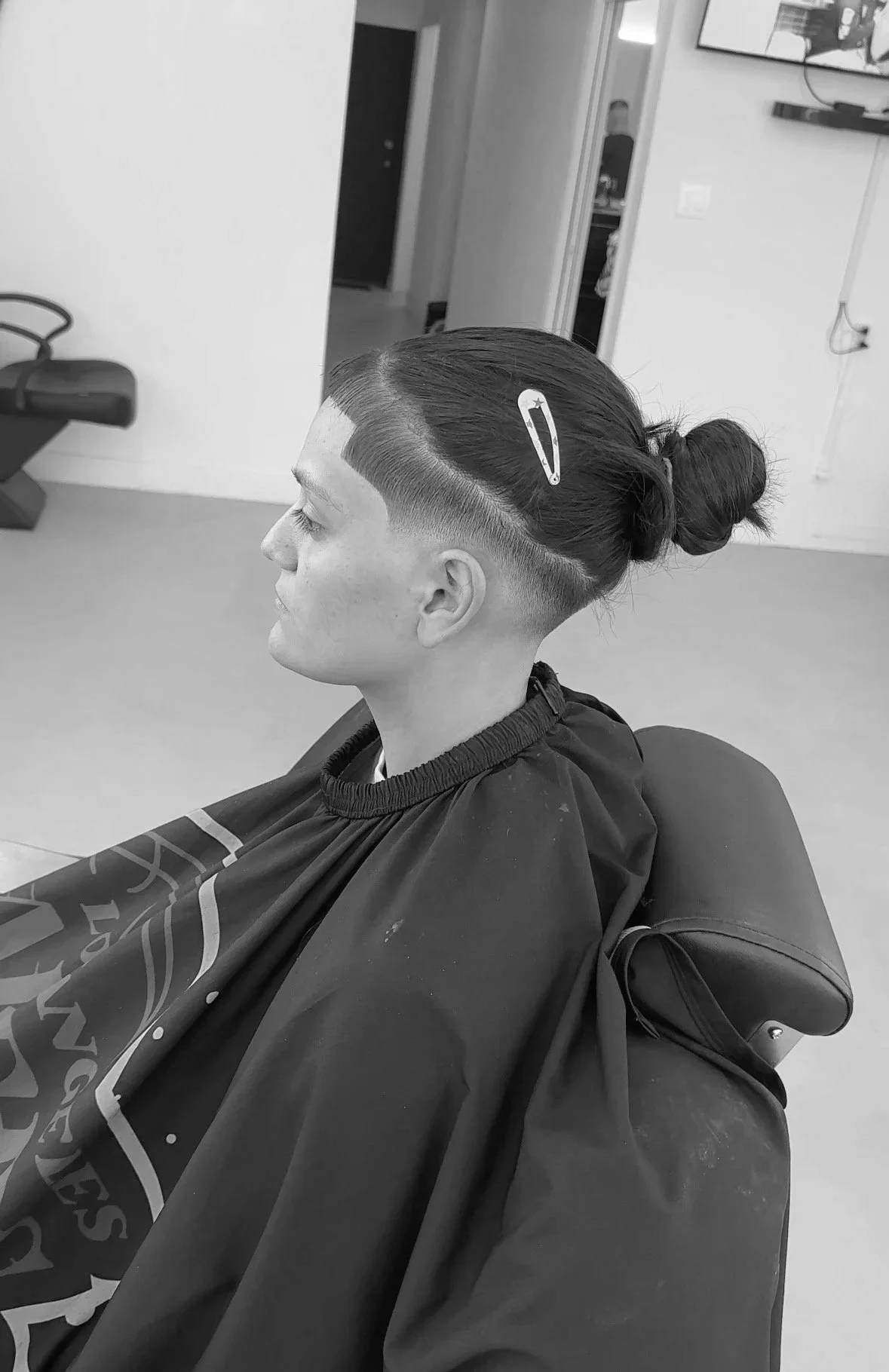 Black & white photo of a woman with a short hairstyle and shaved sides, sitting in a barber's chair with a cape around her shoulders, in a barbershop.
