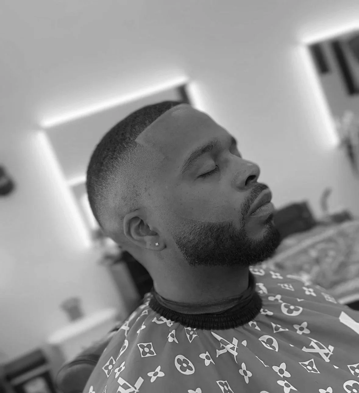 A man with a short hairstyle, beard, and earrings resting with eyes closed in a barber shop chair, wearing a patterned cape.