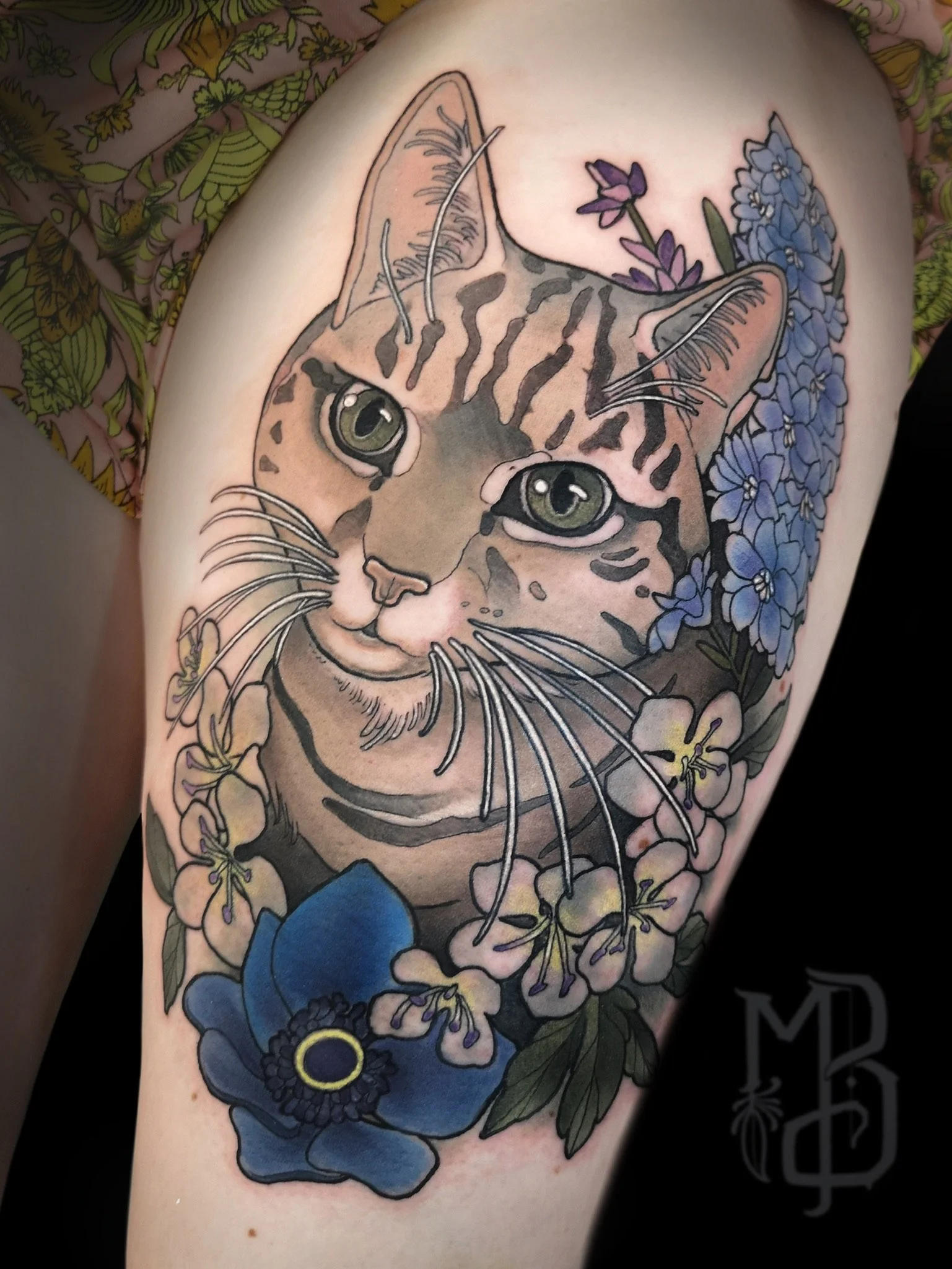 Tattoo of a cat portrait, surrounded by blue, purple, and yellow flowers on a person's arm.