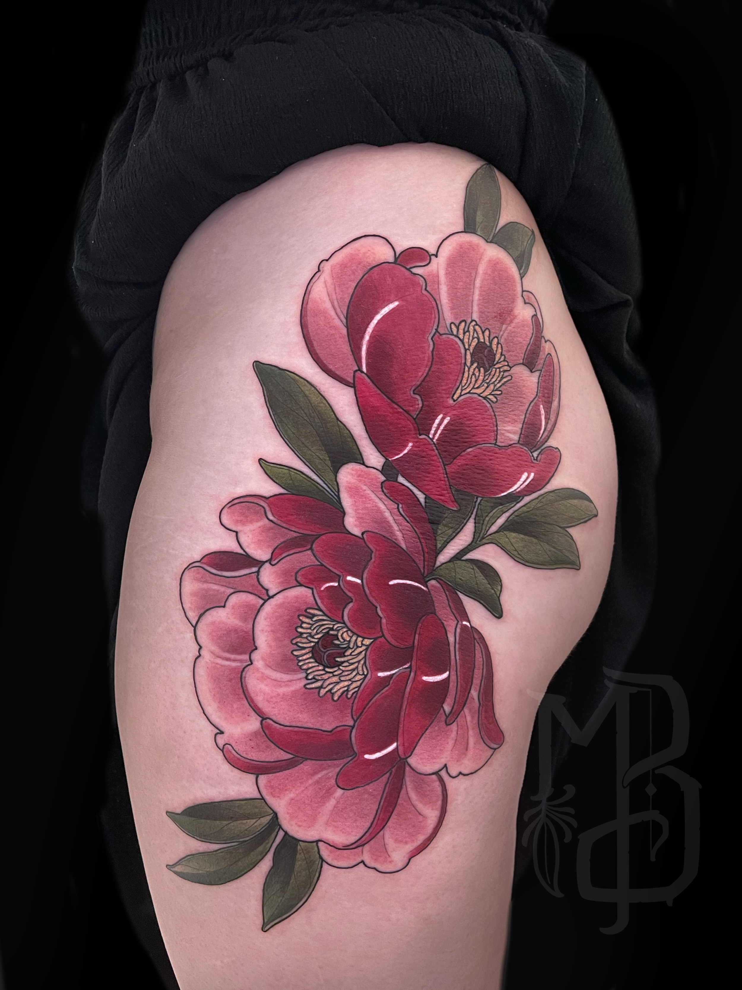 A colorful and feminine neotraditional floral tattoo of large red and pink peony flowers with green leaves on the upper thigh of a person wearing black shorts.