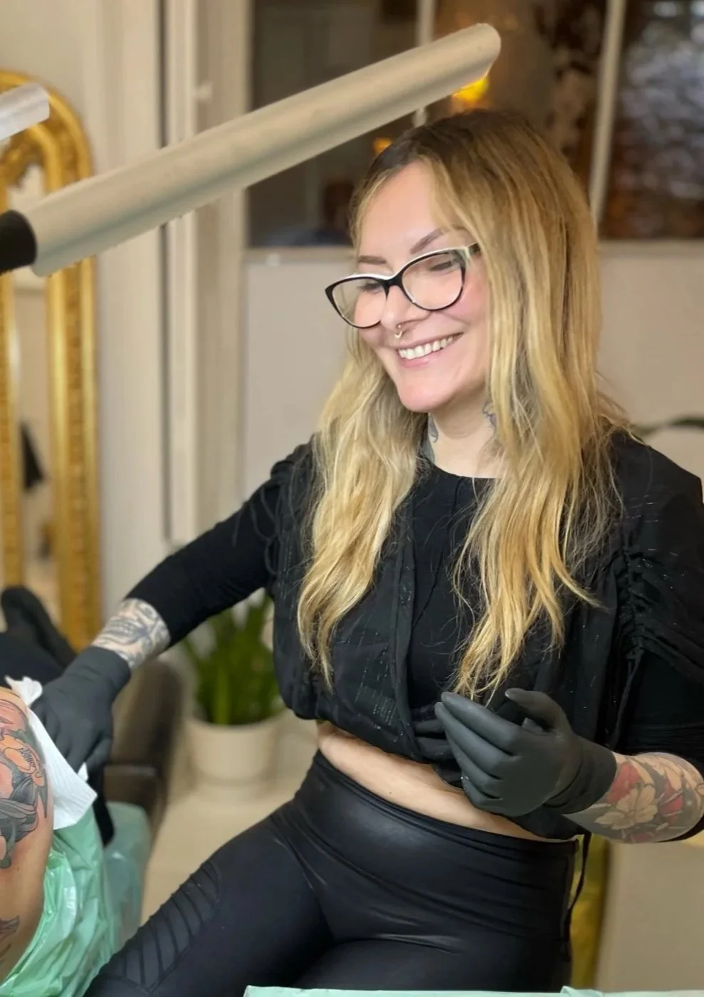 A woman tattooer with long blonde hair, glasses, and tattoos smiling while tattooing a client. She is wearing black gloves and a black top, in a tattoo studio in Dublin City Centre