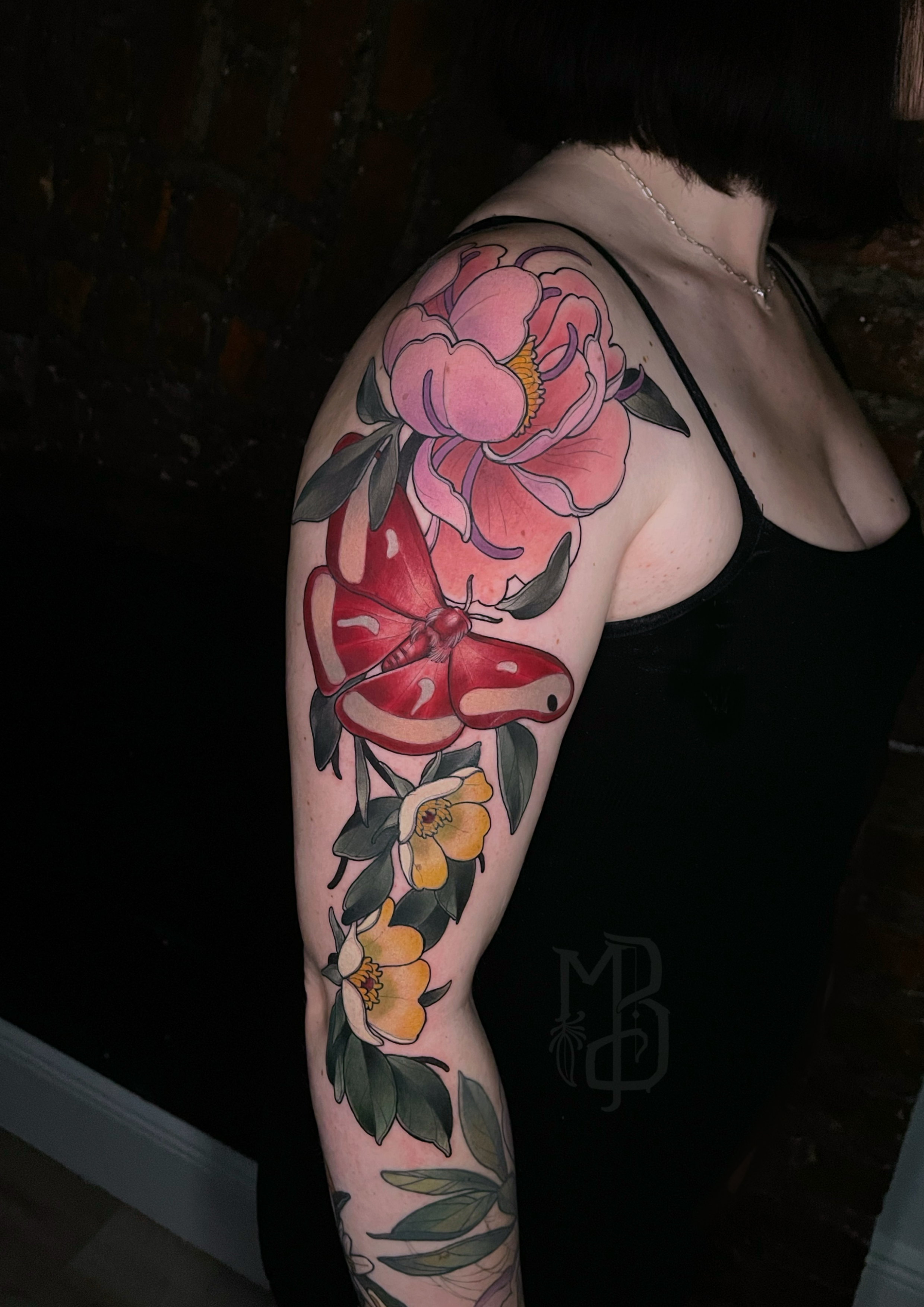 A shoulder and arm neotraditional tattoo of a large pink peony flower, red moth, smaller yellow flowers, and green leaves .