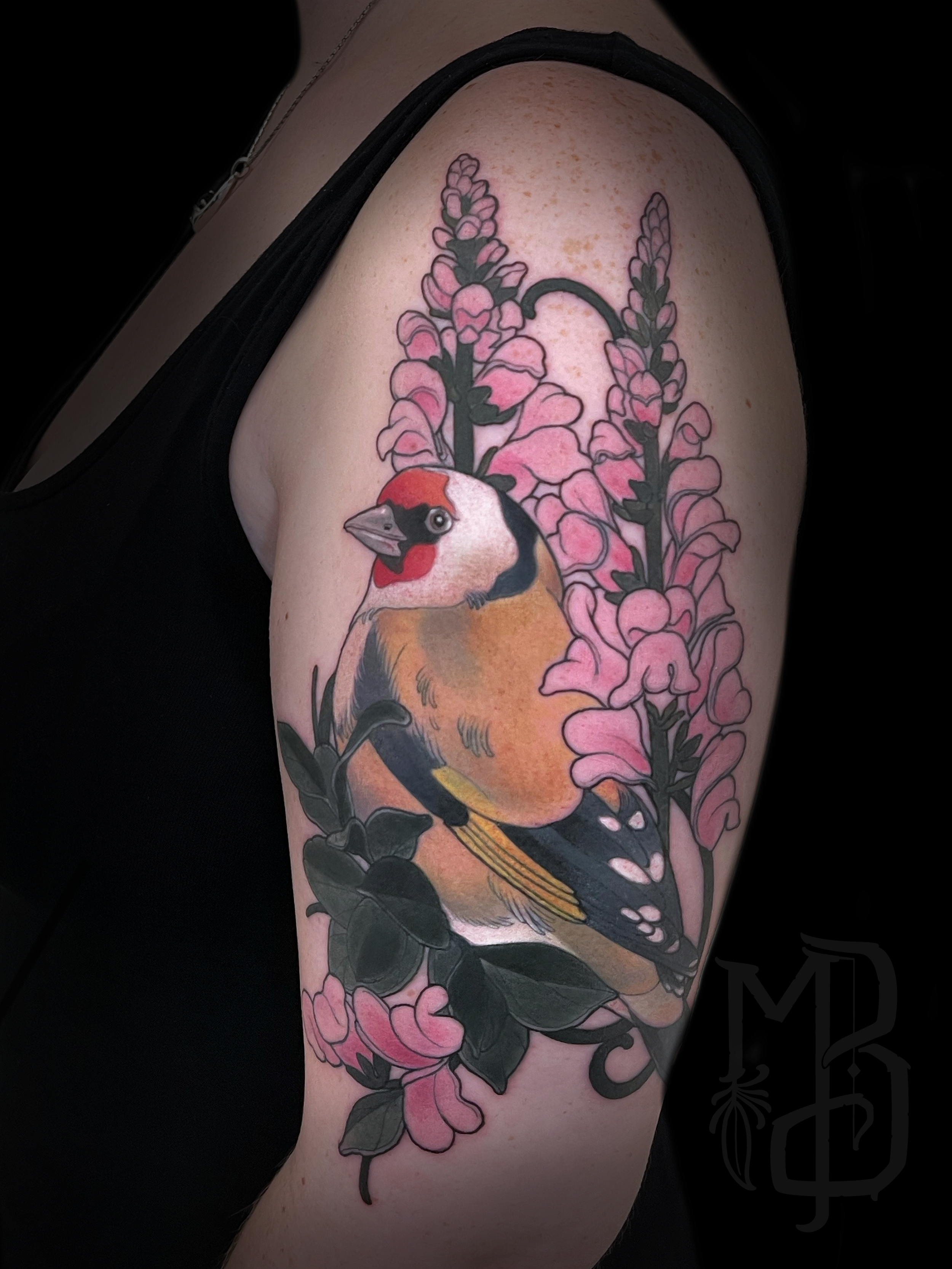 Tattoo of a neotraditional bird, possibly a goldfinch, surrounded by pink flowers and green leaves on upper arm.