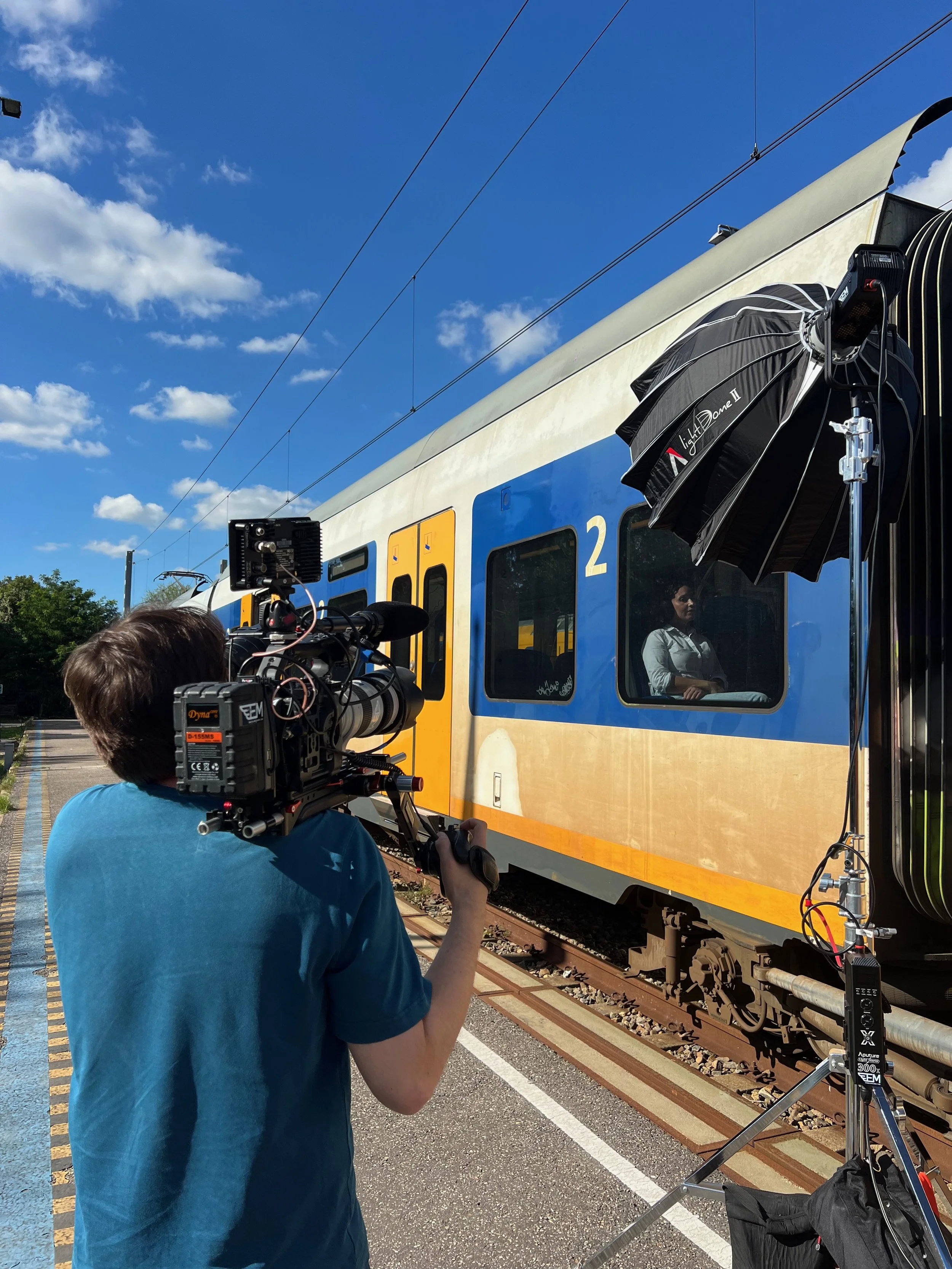 Een filmploeg filmt een blauwe en gele trein terwijl een vrouw in de trein naar de camera kijkt, met een groot verlichtingsapparaat en filmapparatuur op de voorgrond.