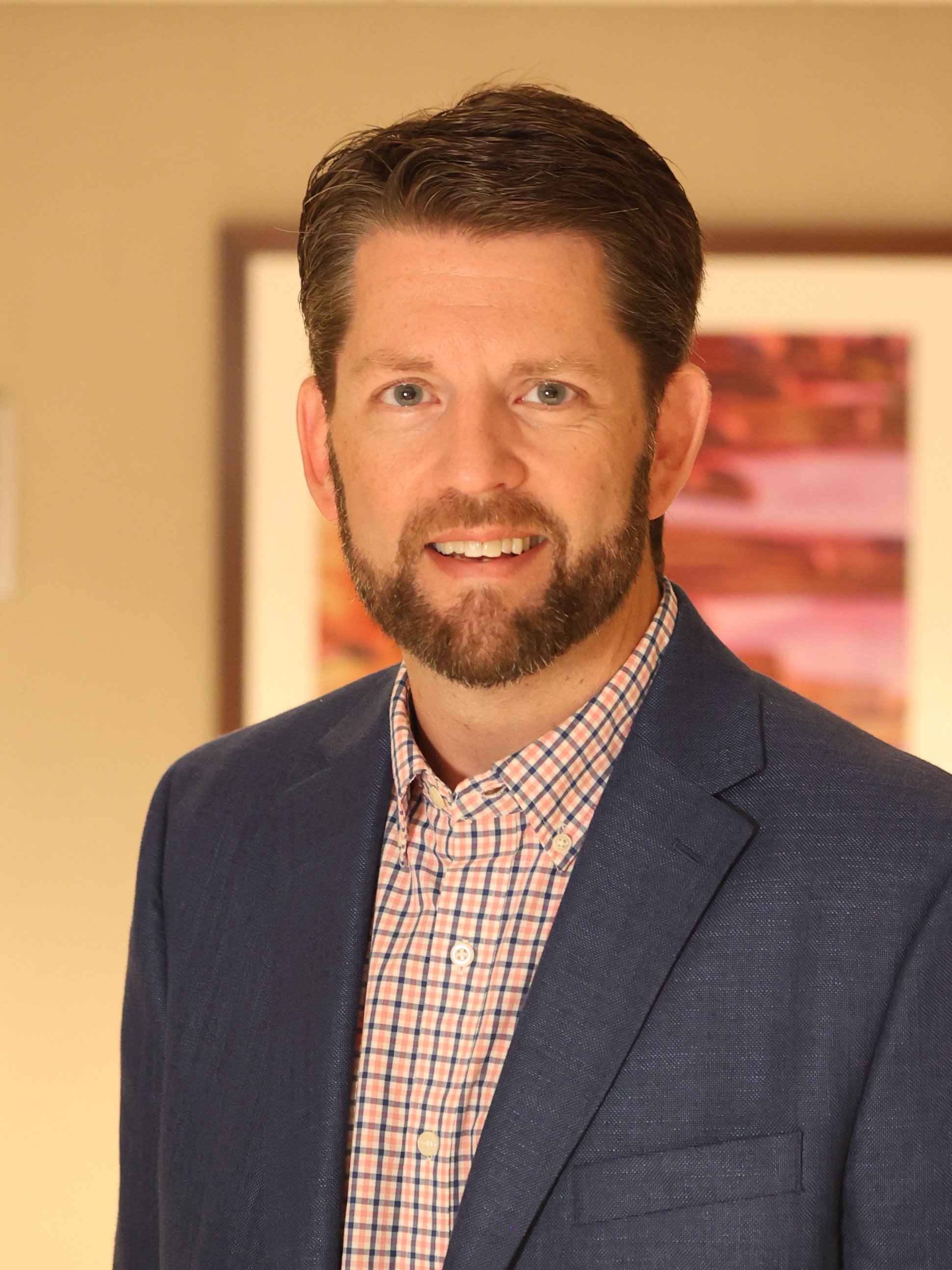 Headshot of a man with brown hair, blue eyes, and a beard, wearing a checked shirt and navy blazer.