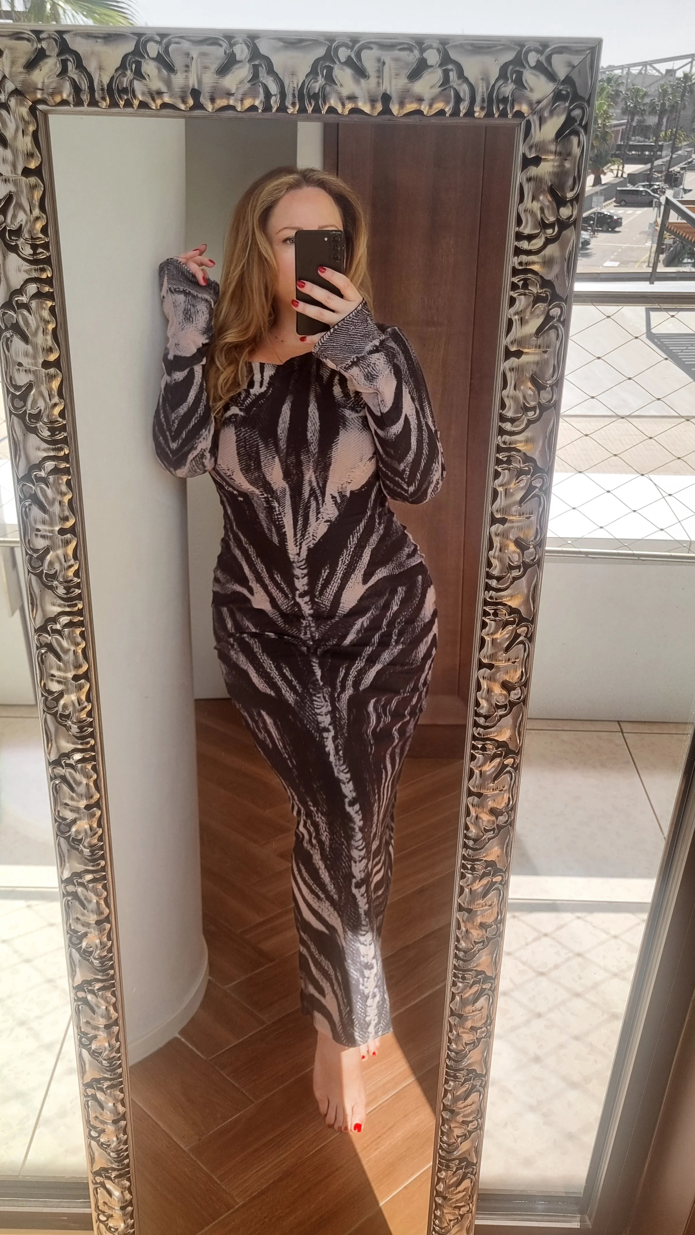 A woman with long, wavy hair takes a mirror selfie, wearing a form-fitting, black and beige patterned dress, standing barefoot in a room with wooden flooring and a decorative mirror frame.