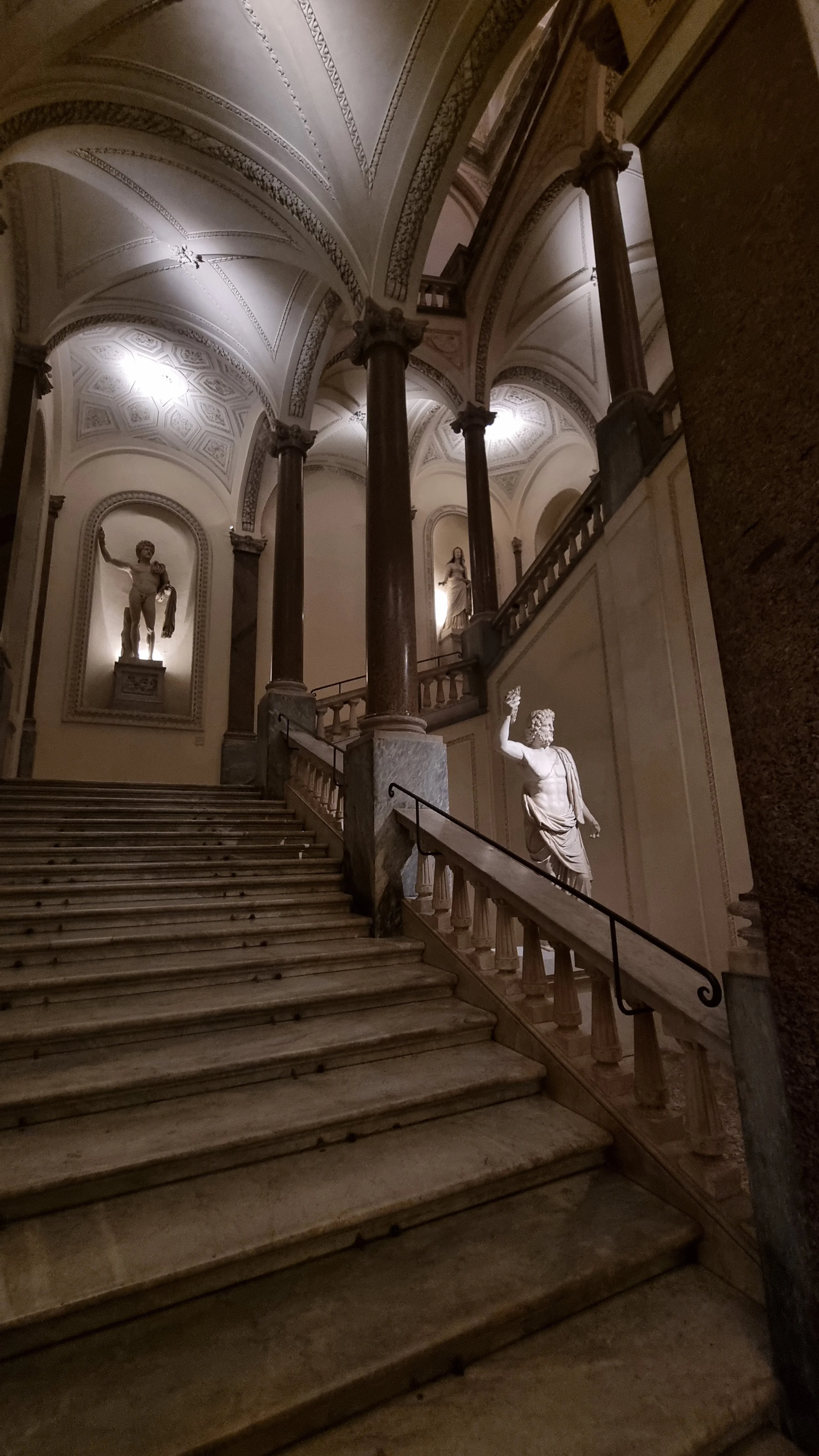 Marble staircase leading up to an ornate hall with classical sculptures and decorated arches and ceiling.