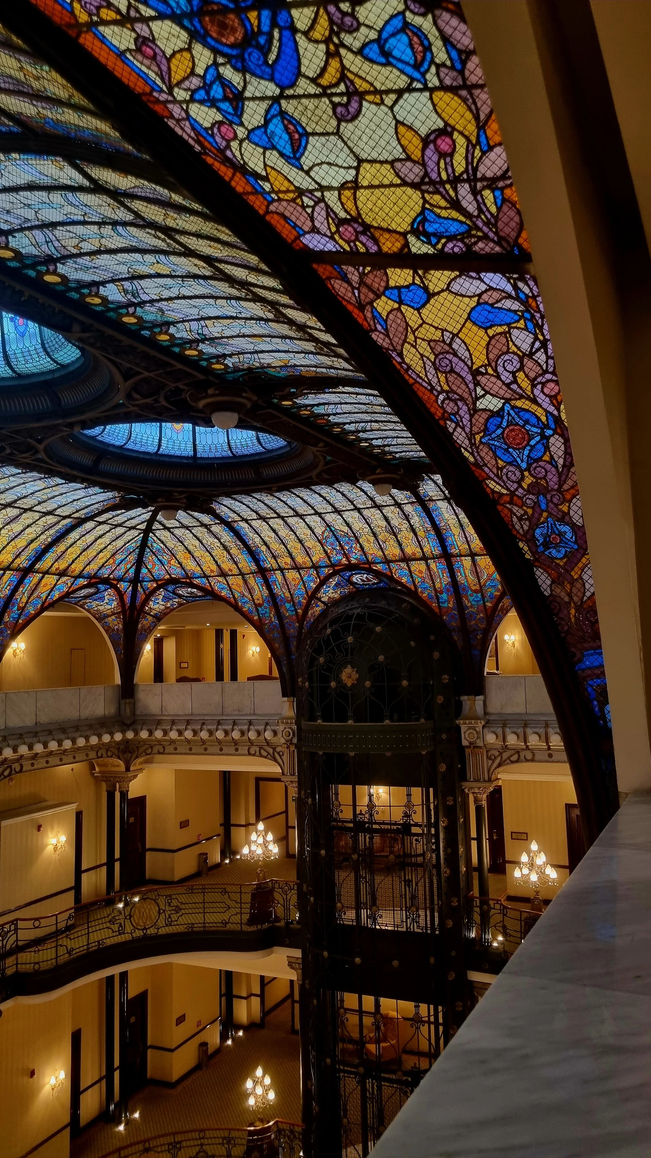 Interior view of a hotel lobby with a large stained glass ceiling featuring intricate floral and geometric designs in blue, purple, yellow, and orange colors, a decorative elevator with ornate ironwork, and elegant chandeliers and wall sconces illumi