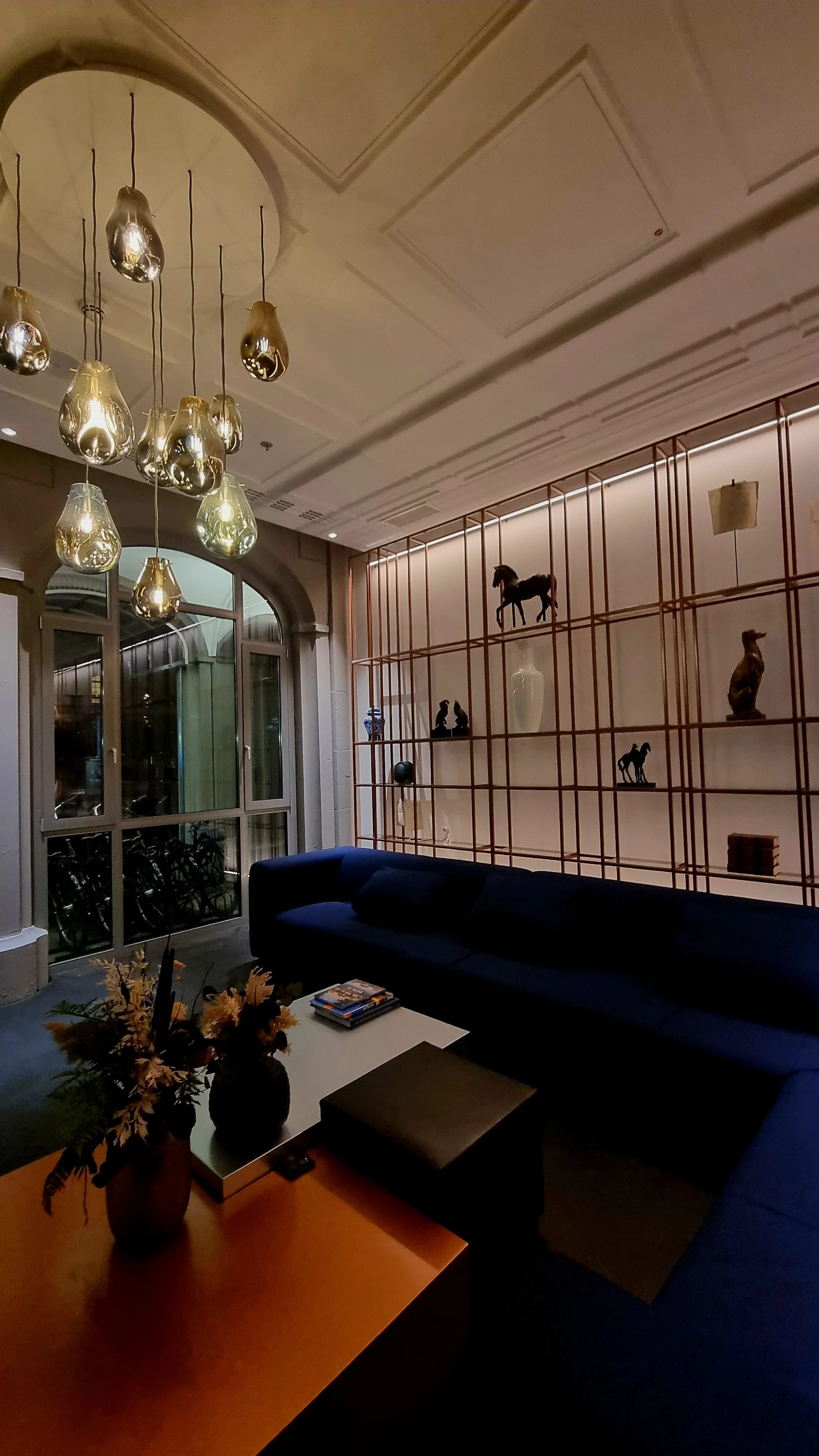 A cozy living room with a blue sofa, a white coffee table with magazines, a floral arrangement in a vase, a large window with a metal frame, a decorative wooden shelf with animal figurines and a large vase, leaf-patterned hanging pendant lights, and 