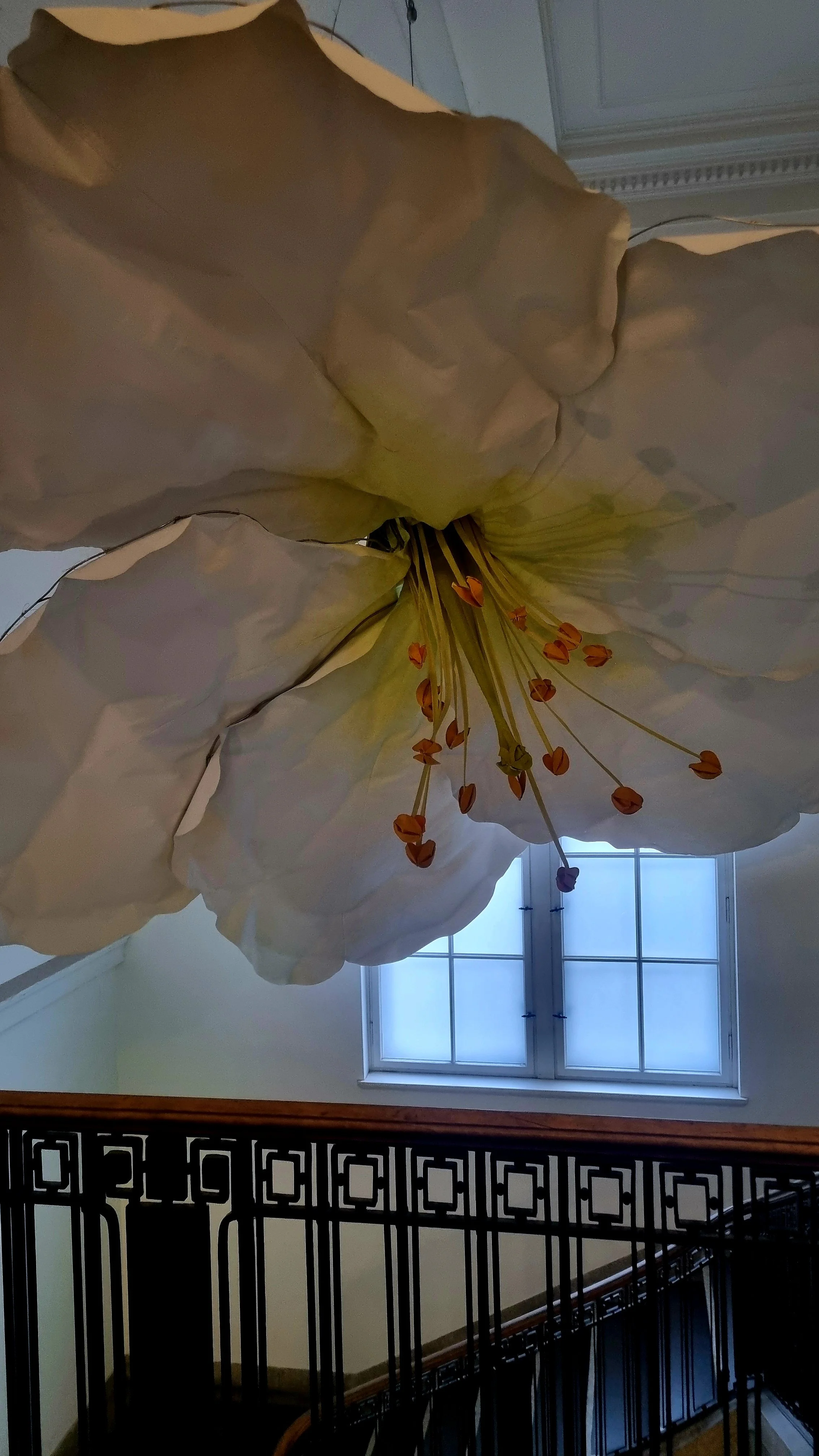 Large artificial white flower with orange stamen hanging from the ceiling near a window, viewed from below, with a black decorative railing in the foreground.