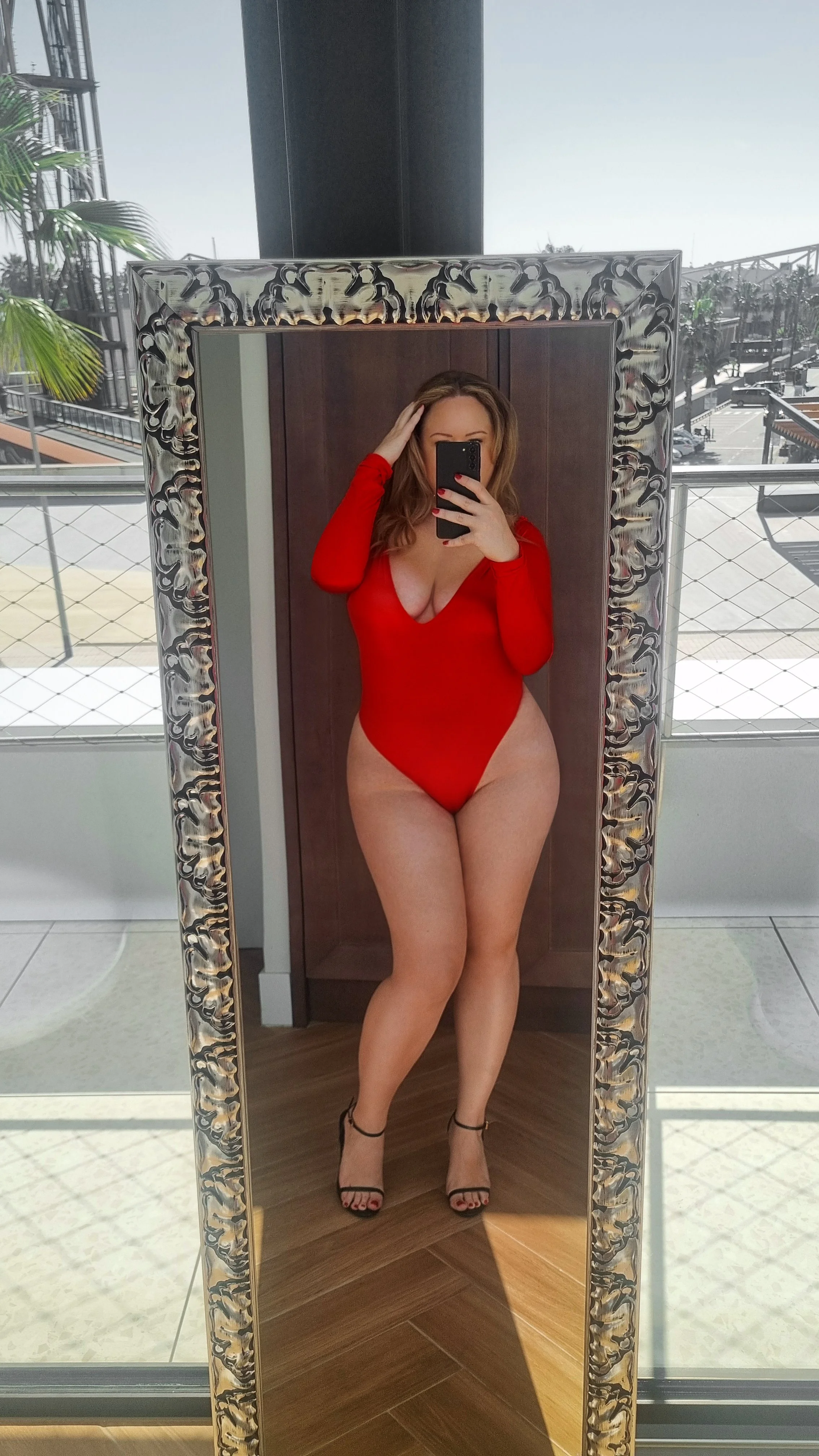Woman in a red long-sleeve swimsuit taking a mirror selfie indoors, with high-heeled sandals, standing on a wooden floor.