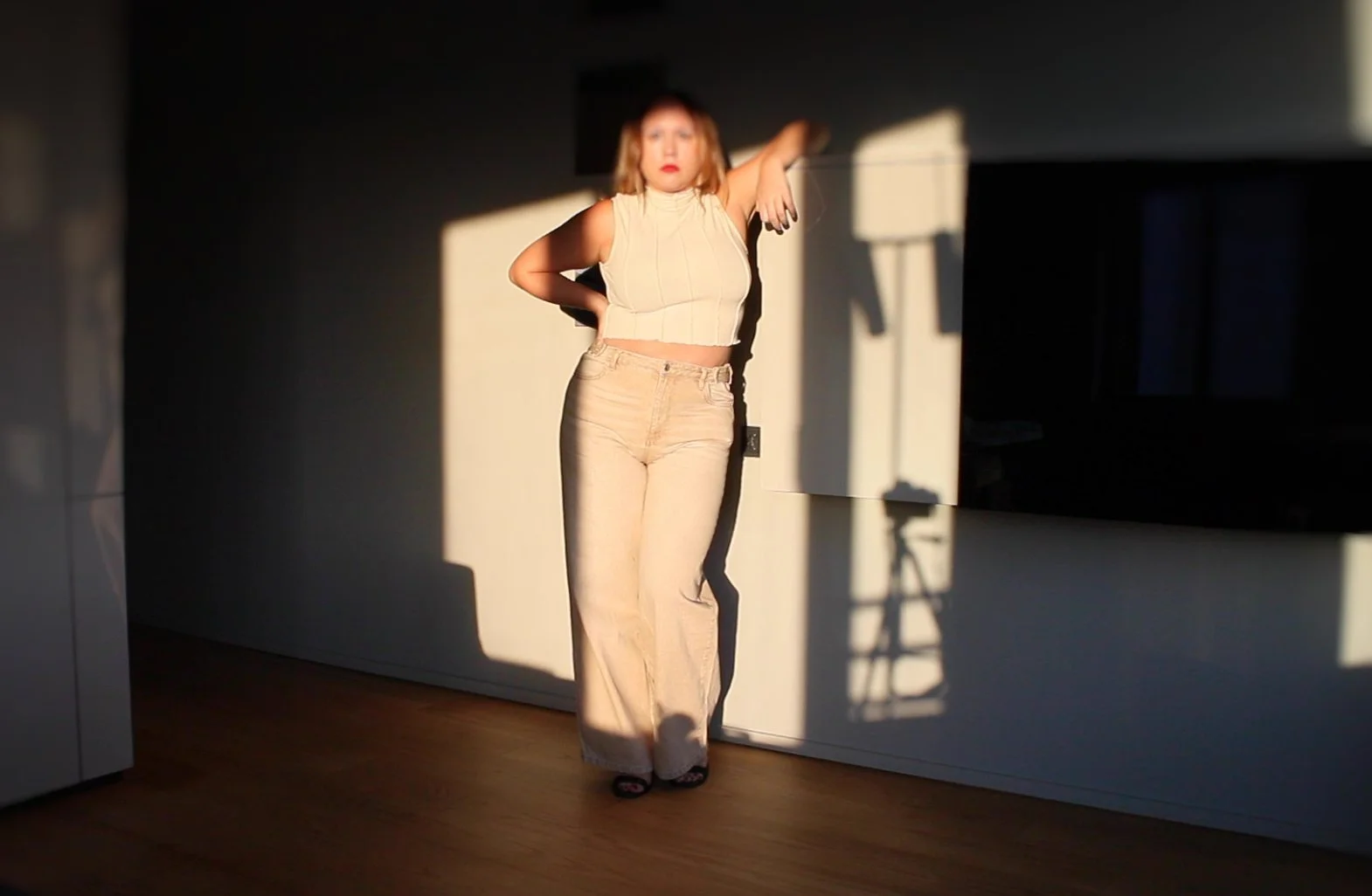 A woman standing indoors in front of a wall, illuminated by sunlight, with her left arm resting on her head and her right arm on her hip, wearing a sleeveless beige top and light-colored pants.