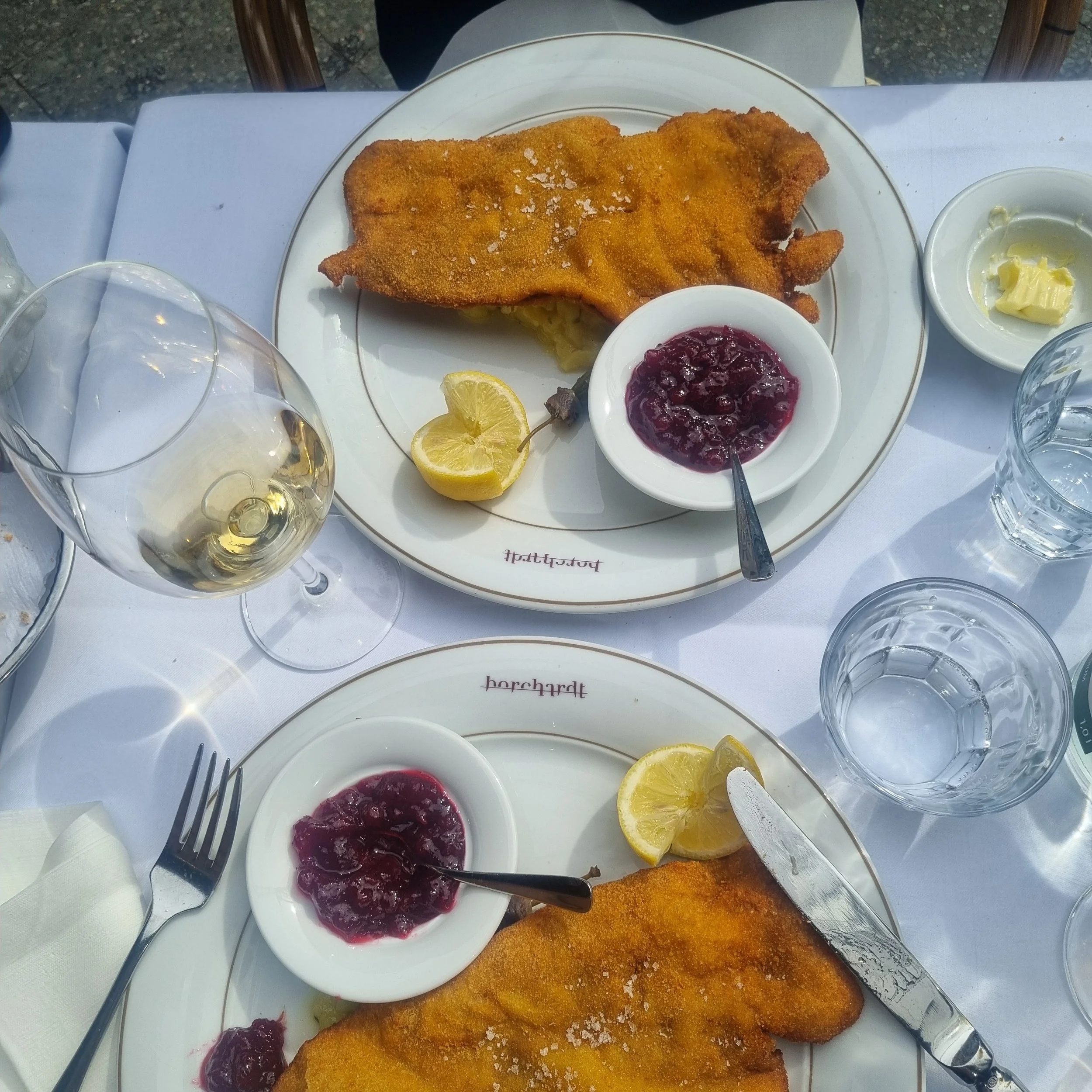 A plate with a large, breaded fried fish fillet, lemon slices, a small bowl of lingonberry jam, and a side of butter. Surrounding the plate are empty glasses and utensils on a white tablecloth.