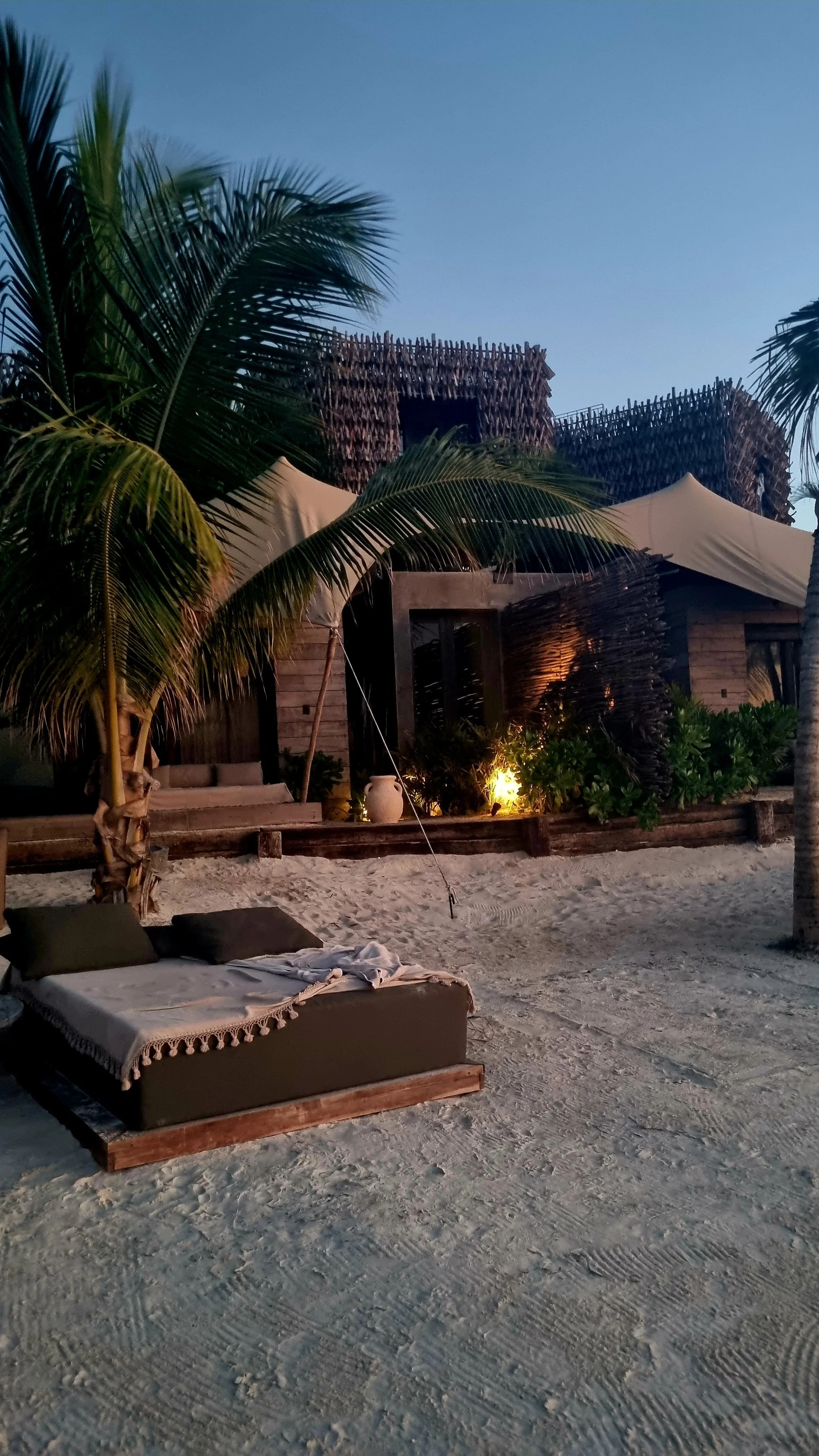 Beachside cabana with palm trees, lounge chair with cushion, sandy ground, and ambient lighting in the evening.