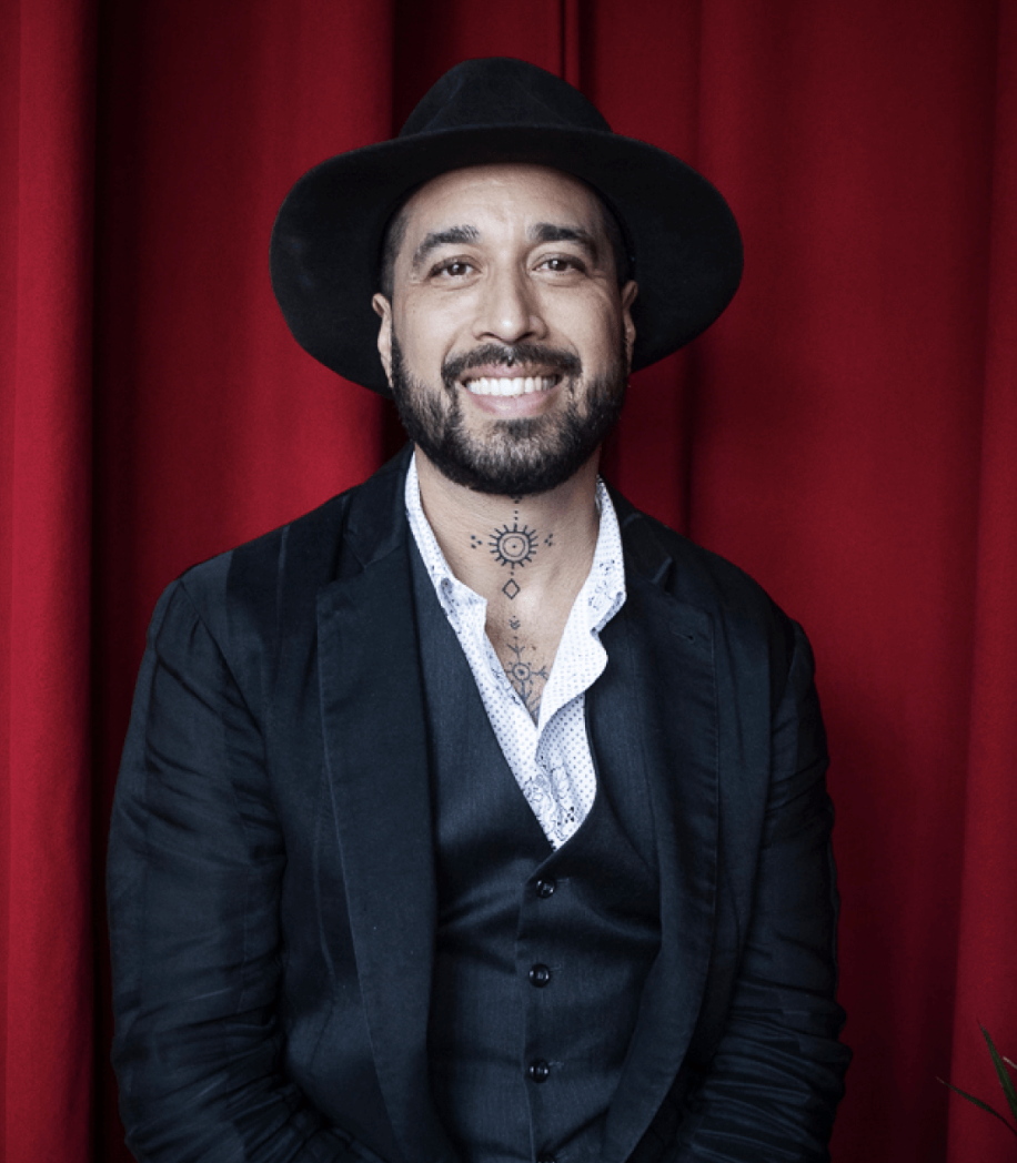 A man with a beard and tattoos wearing a black hat, black jacket, and a white shirt, smiling in front of red curtains.