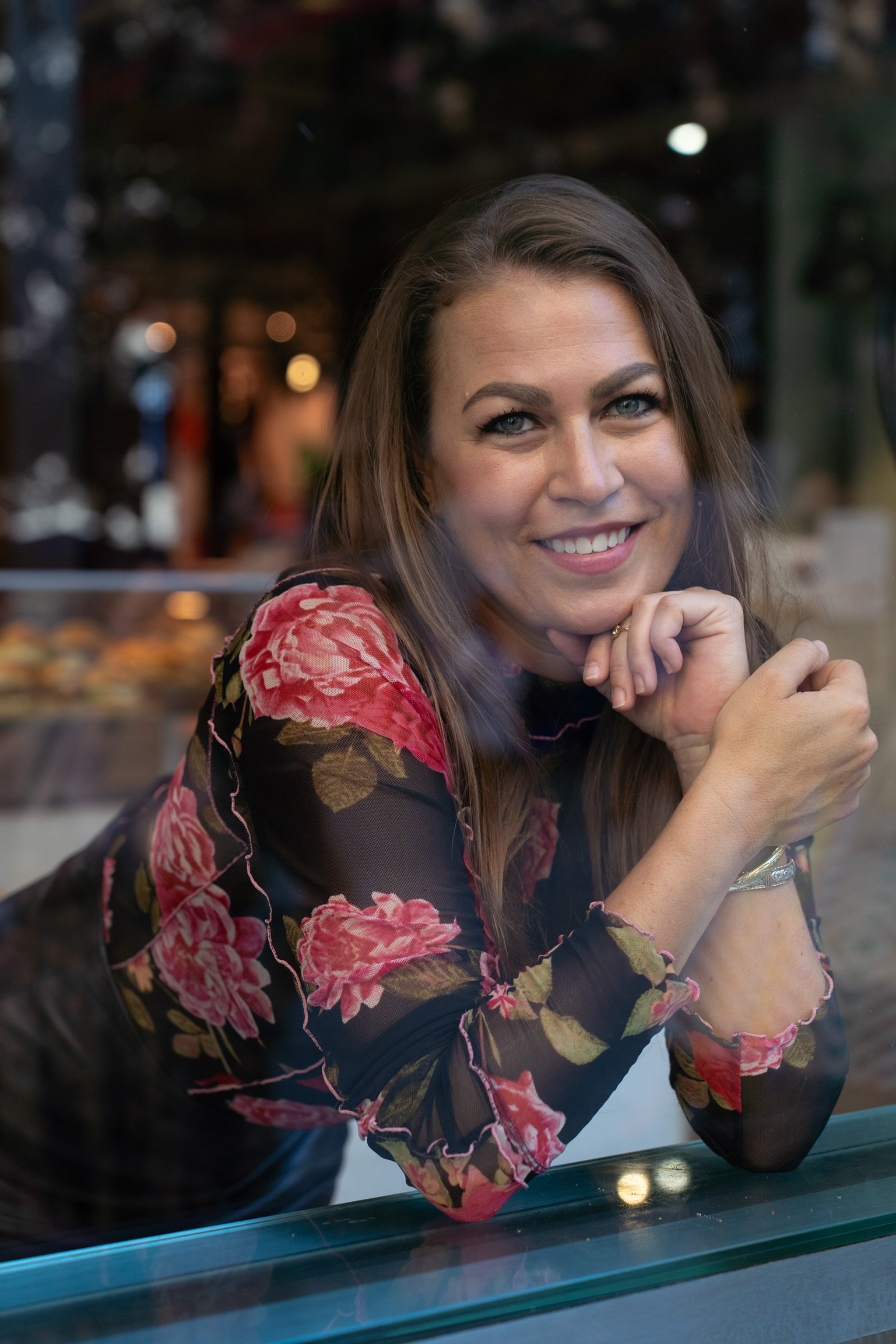 A woman with long brown hair, wearing a black top with pink and red floral print, smiling and resting her chin on her hand, sitting at a table in a dimly lit indoor setting.