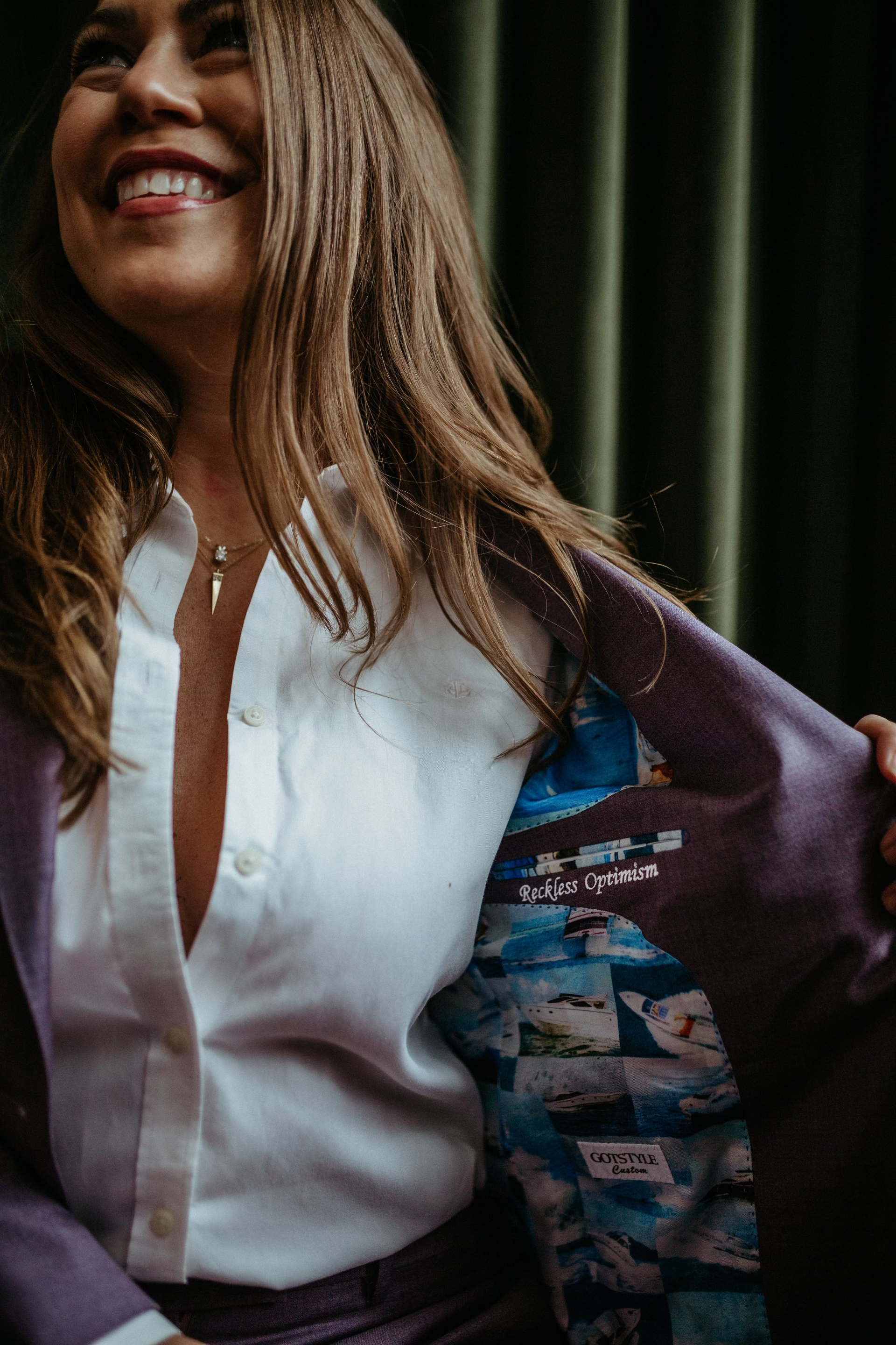 A woman smiling, wearing a white button-up shirt, and opening a purple blazer with a colorful lining that has images of boats and the words 'Reckless Optimism' and 'Goodtime.'