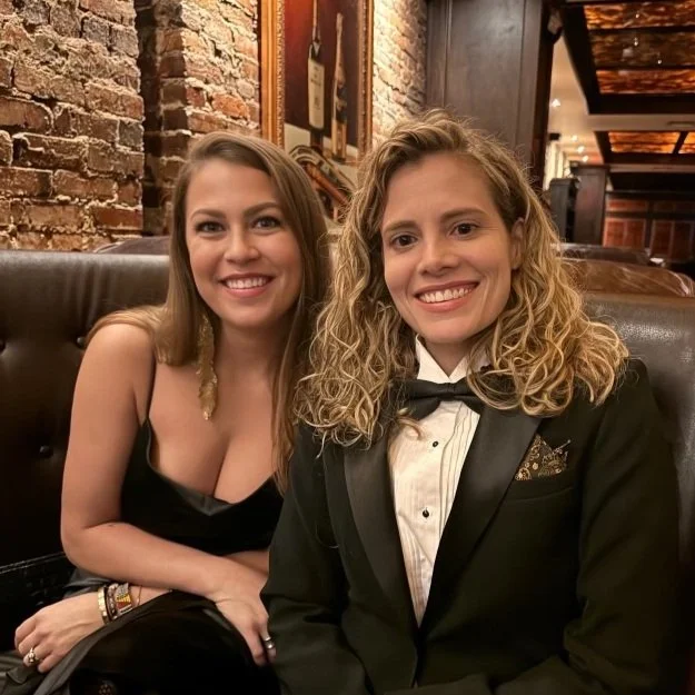 Two women sitting together in a cozy restaurant, smiling at the camera, one in a black dress and the other in a tuxedo, with a rustic brick wall and framed pictures in the background.