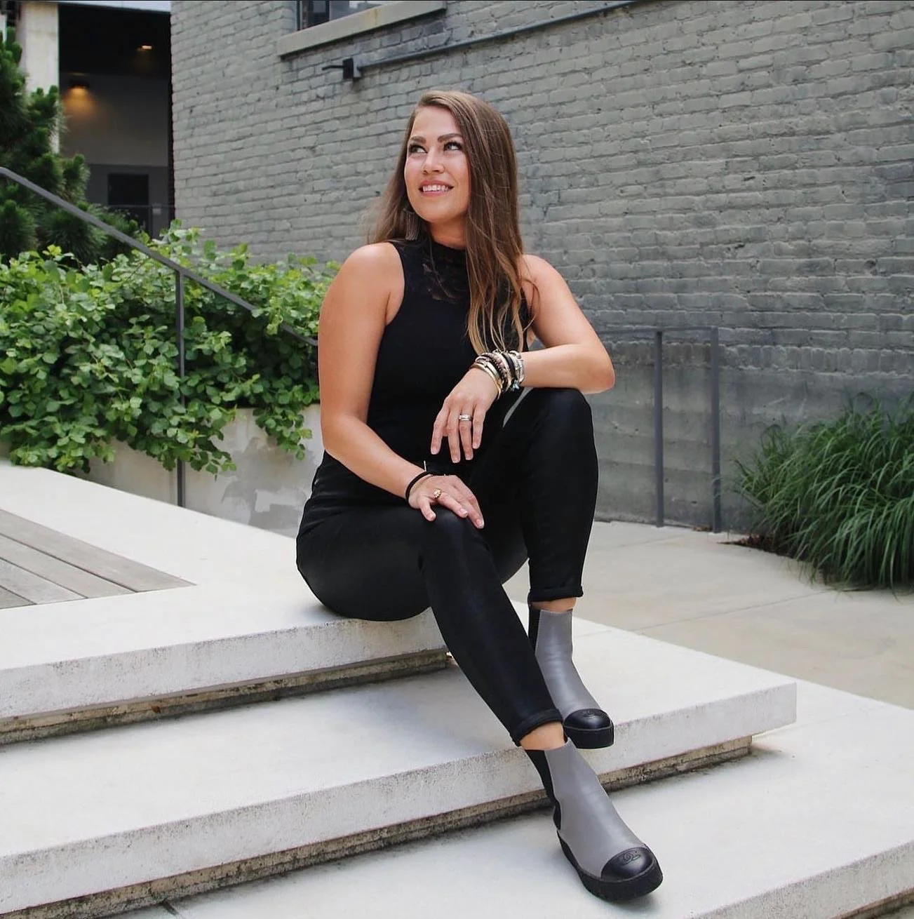 A woman sitting on concrete steps outdoors, wearing a black sleeveless top, black pants, and gray ankle boots, with jewelry on her wrist and fingers, smiling and looking to her left against a gray brick wall with greenery.