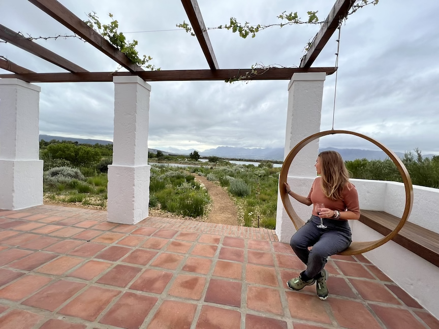 A woman sitting on a round hanging swing with a wine glass, on a terracotta tiled patio, overlooking a lush green landscape with a dirt path and mountains in the distance, under a cloudy sky.