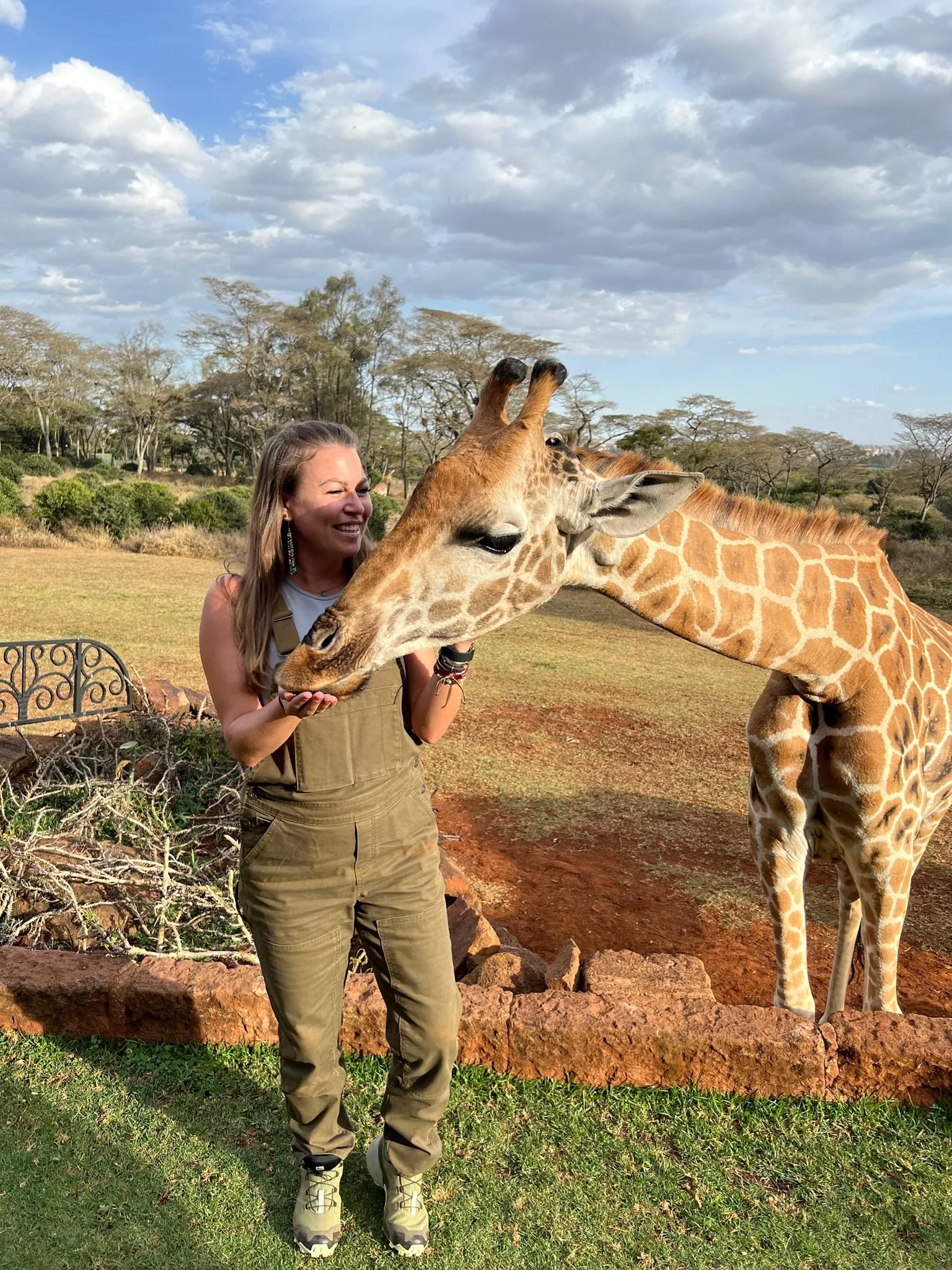 A woman in khaki overalls offering food to a giraffe at a safari park, with trees and a cloudy sky in the background.