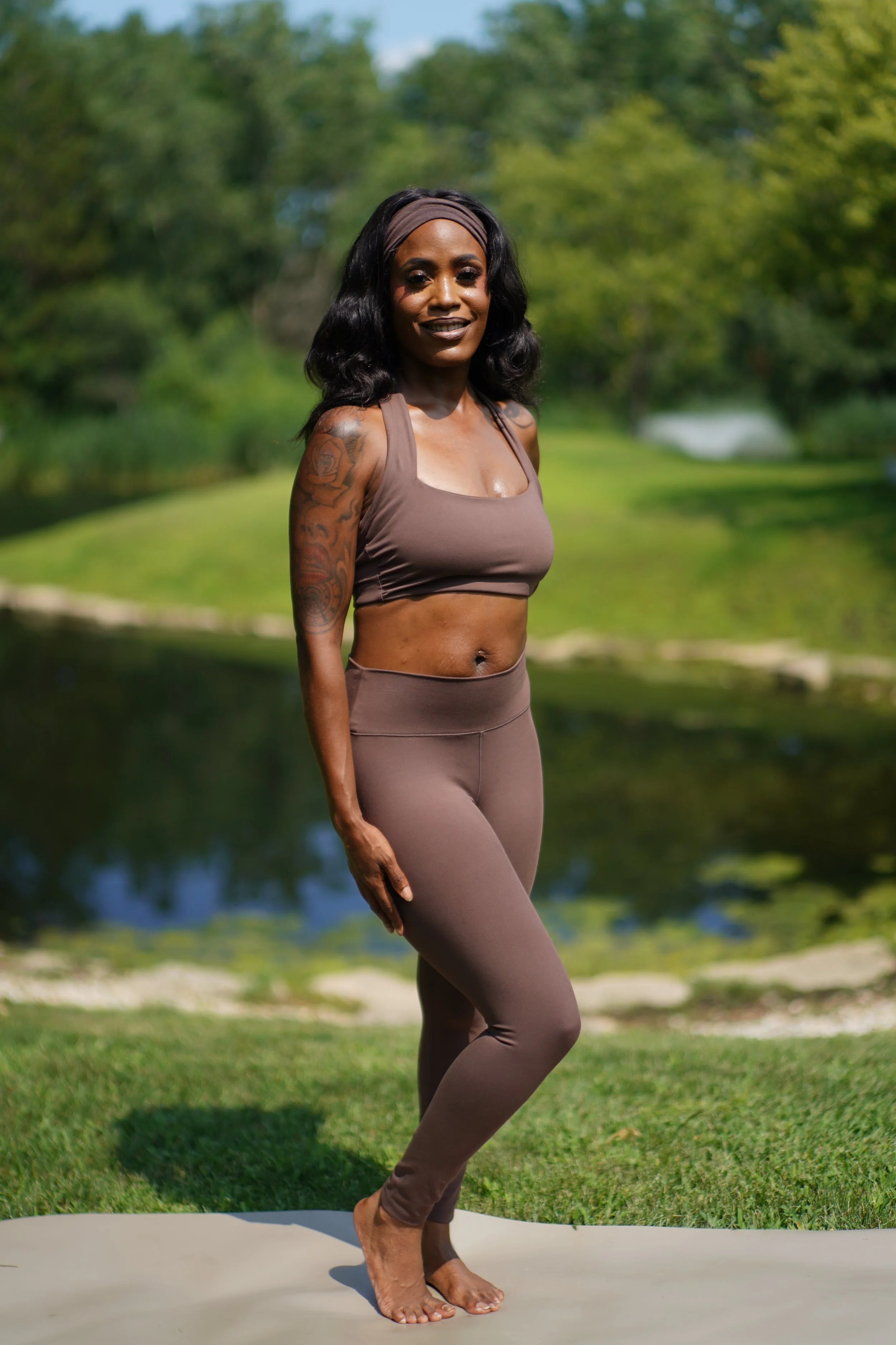 A woman practicing yoga outdoors on a mat near a lake with green trees in the background.
