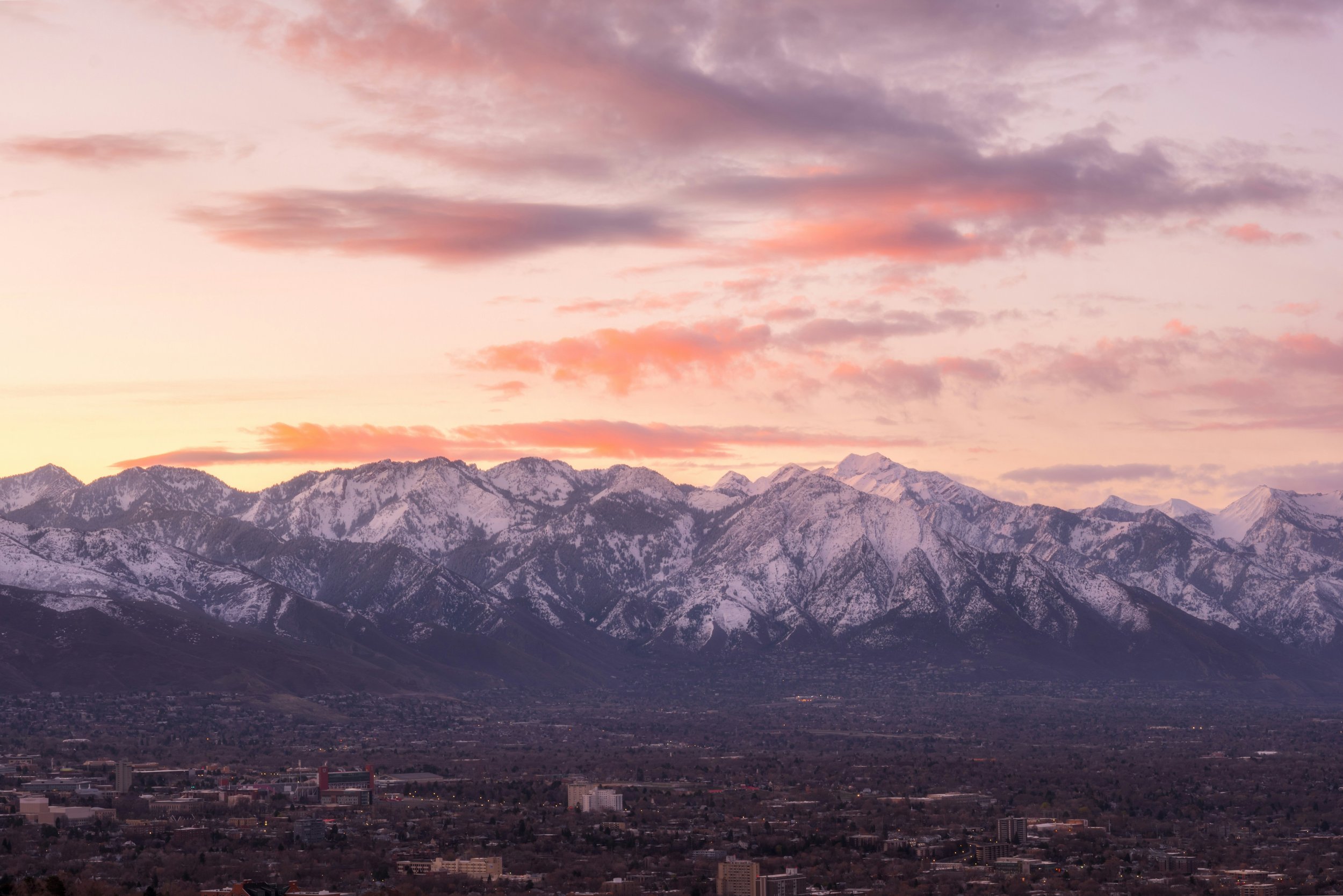Sunset over snow-capped mountains with a pink and purple sky, overlooking a city below.