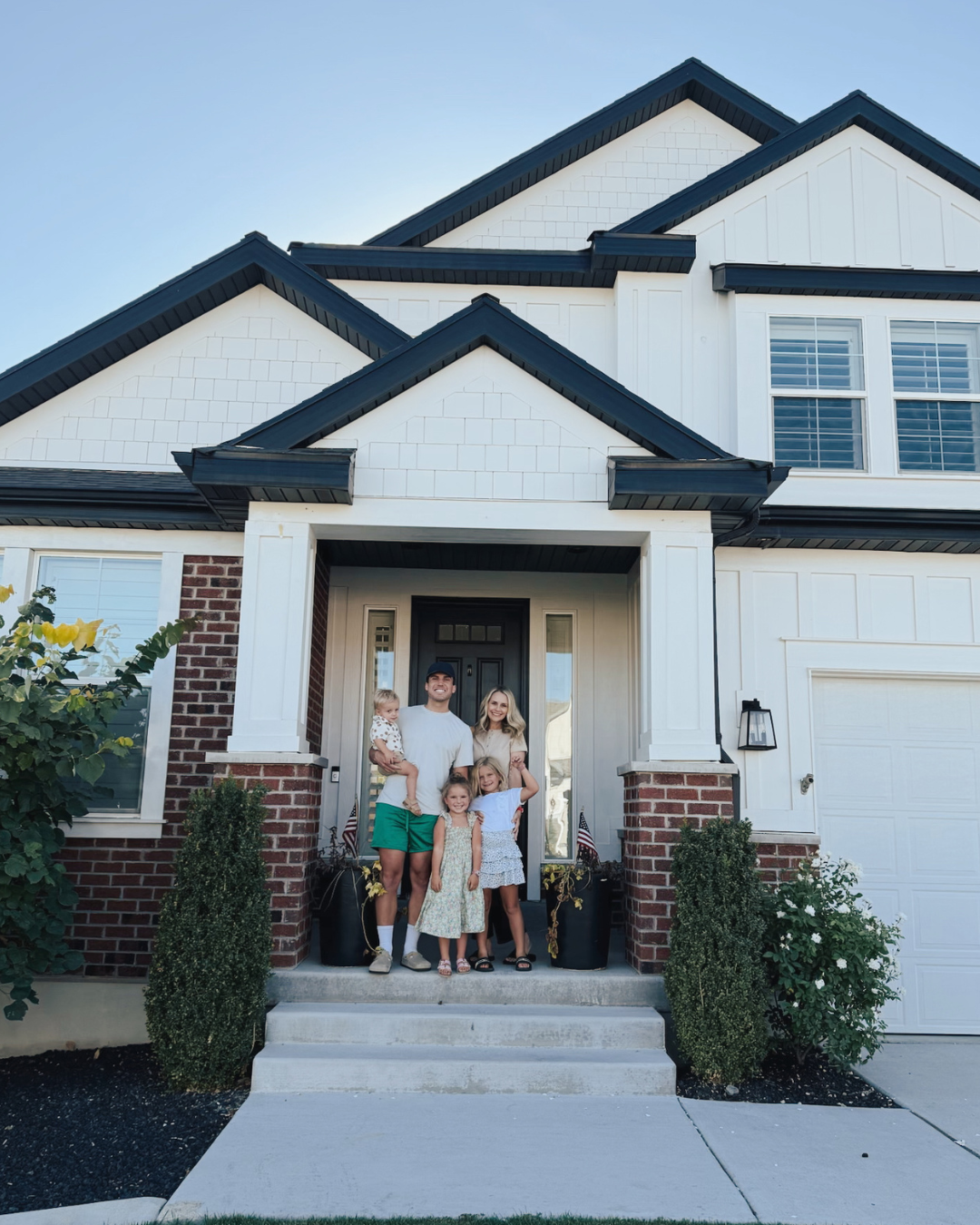 A family standing on the front porch of a modern two-story house with white siding and brick accents, smiling at the camera.