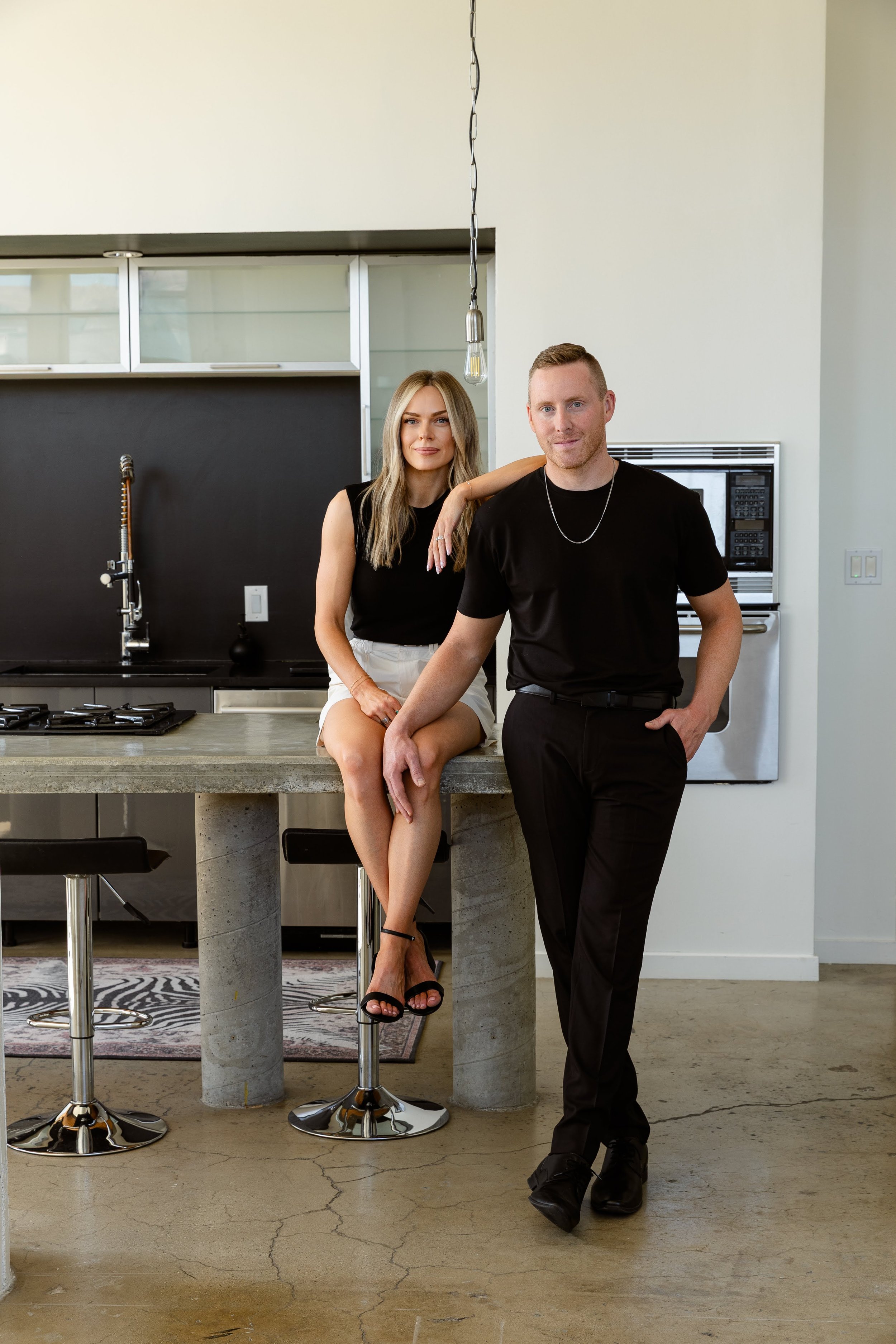 A man and a woman in a modern kitchen with black and white decor. The woman is sitting on the kitchen counter with one leg crossed, and the man is standing beside her with one hand in his pocket. Both are dressed in black tops and dark pants.