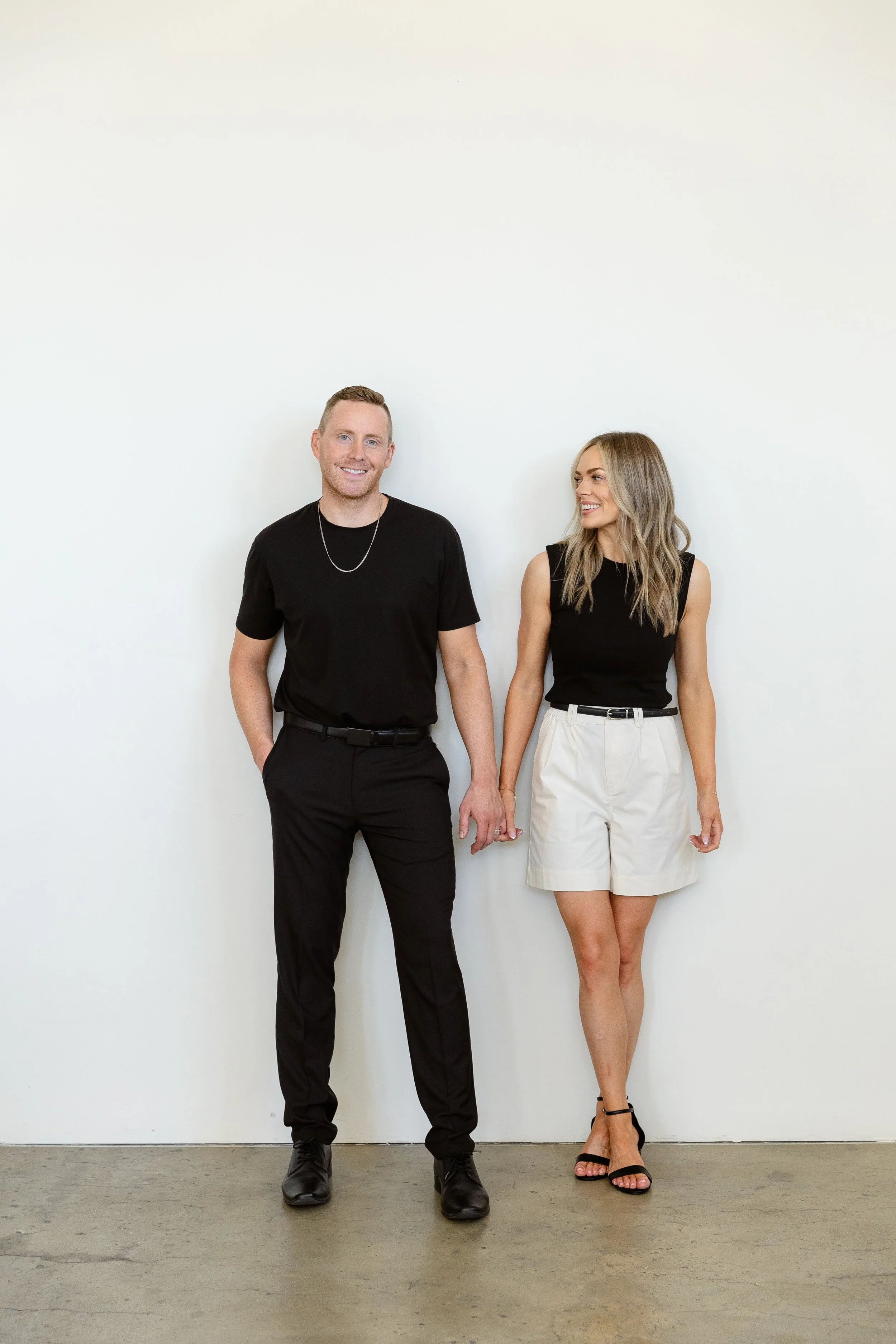 A man and a woman holding hands and standing against a plain white wall, both dressed in black and white clothing. The man is wearing black pants and a black t-shirt, while the woman is wearing a black sleeveless top and beige shorts.