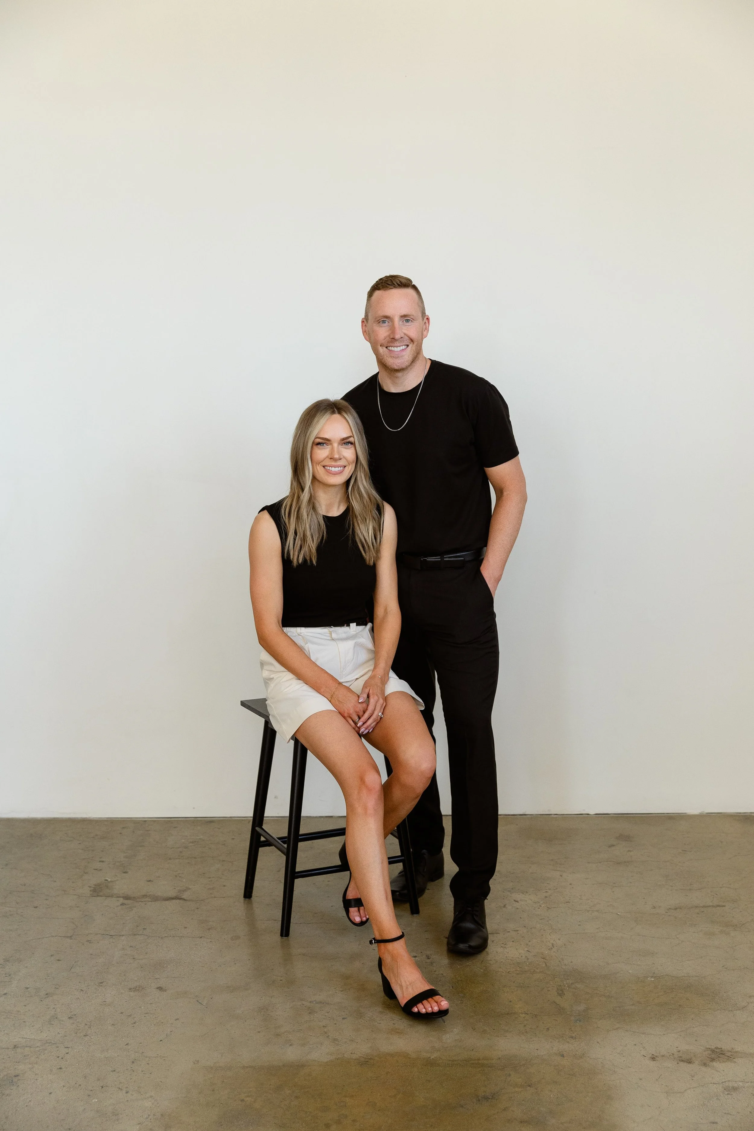A woman sitting on a black stool and a man standing beside her, both smiling against a plain white wall. The woman has long, wavy blonde hair, is wearing a black sleeveless top, white shorts, and black high-heeled sandals. The man has short, light-colored hair, is wearing a black t-shirt, black pants, and black shoes.