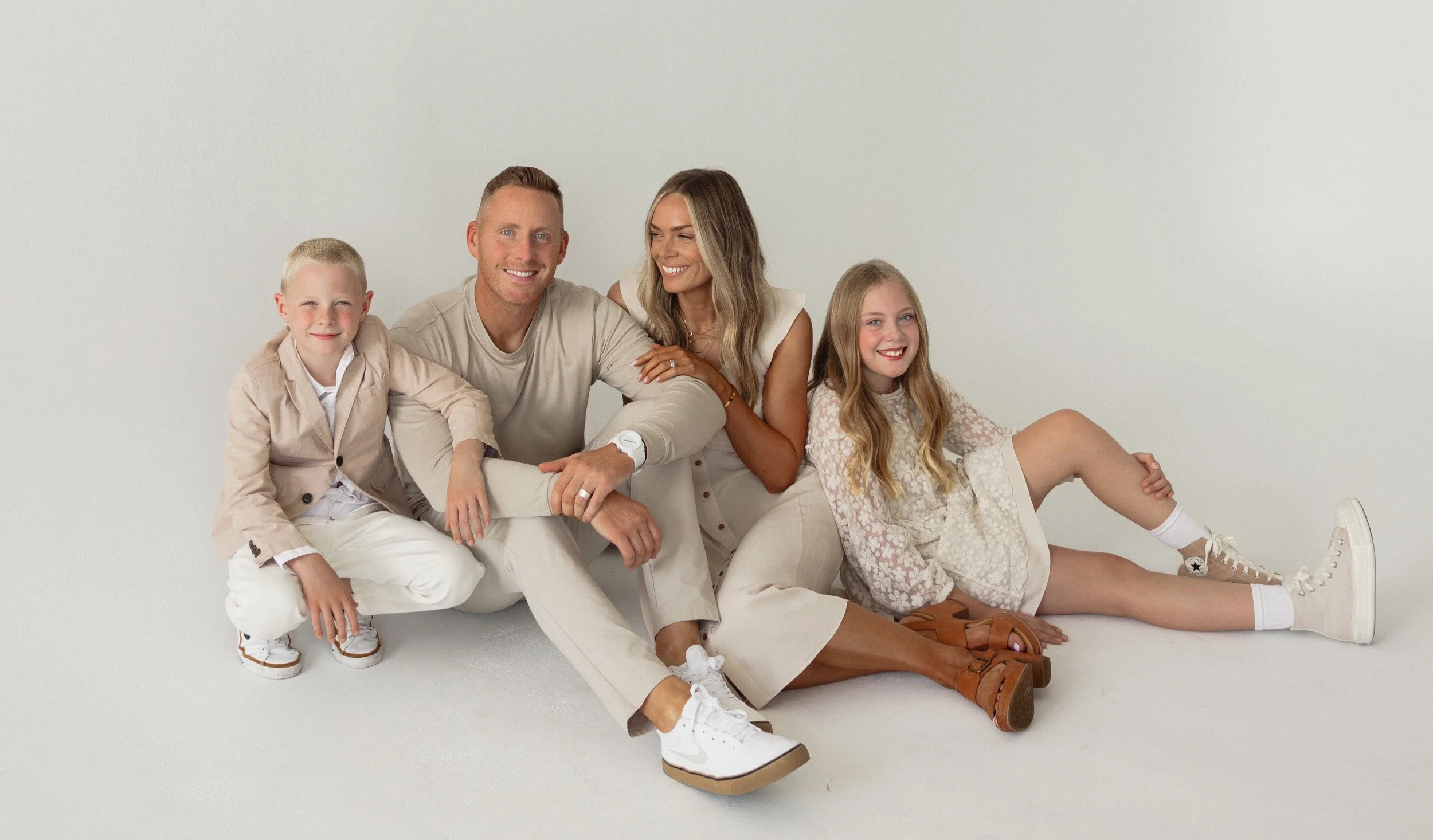 A smiling family of five sitting against a plain white background. The father, mother, and three children are dressed in light-colored casual clothing, sitting and lying on the floor.
