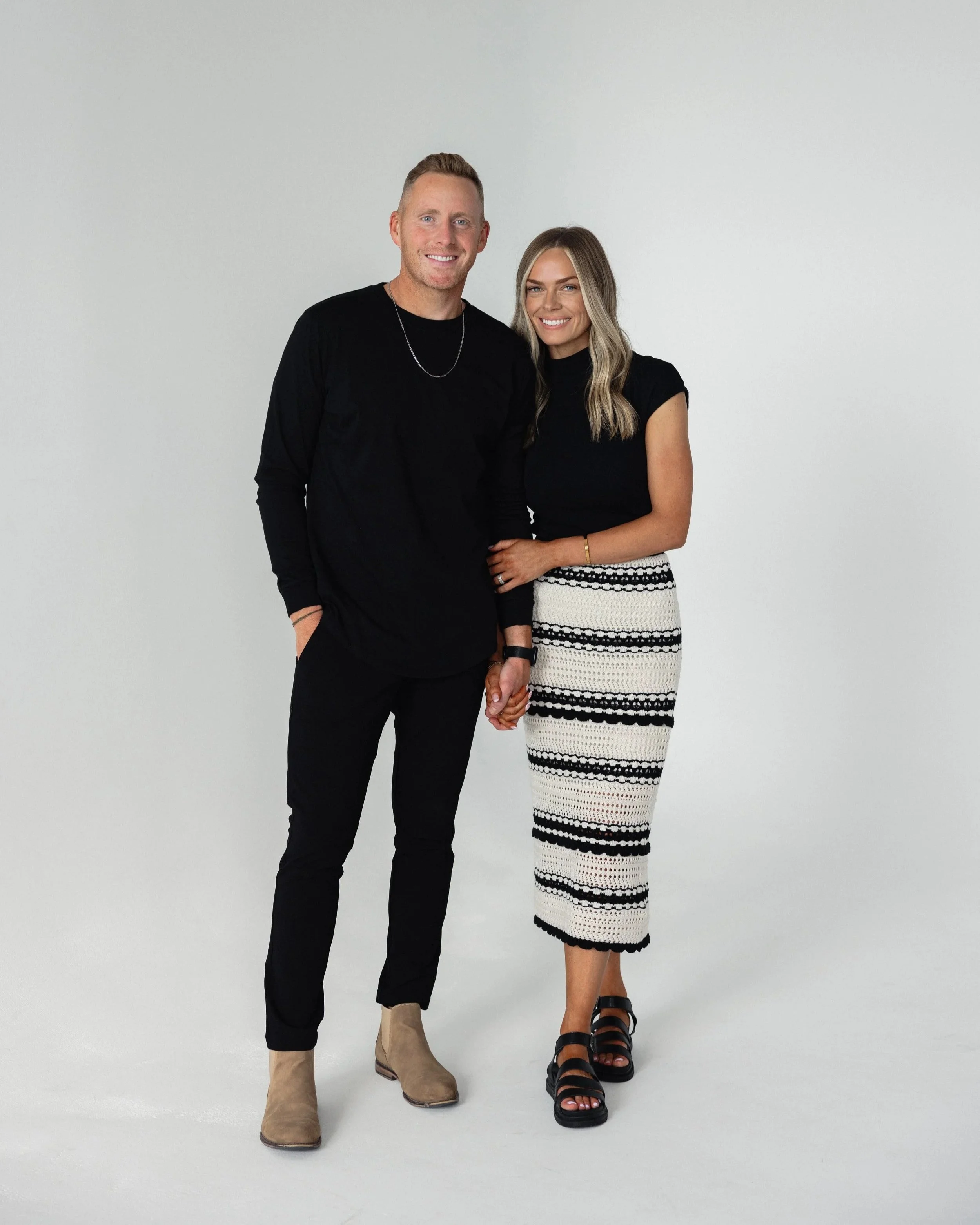 A young man and woman standing together against a plain white background, holding hands, smiling, dressed in black and white outfits.