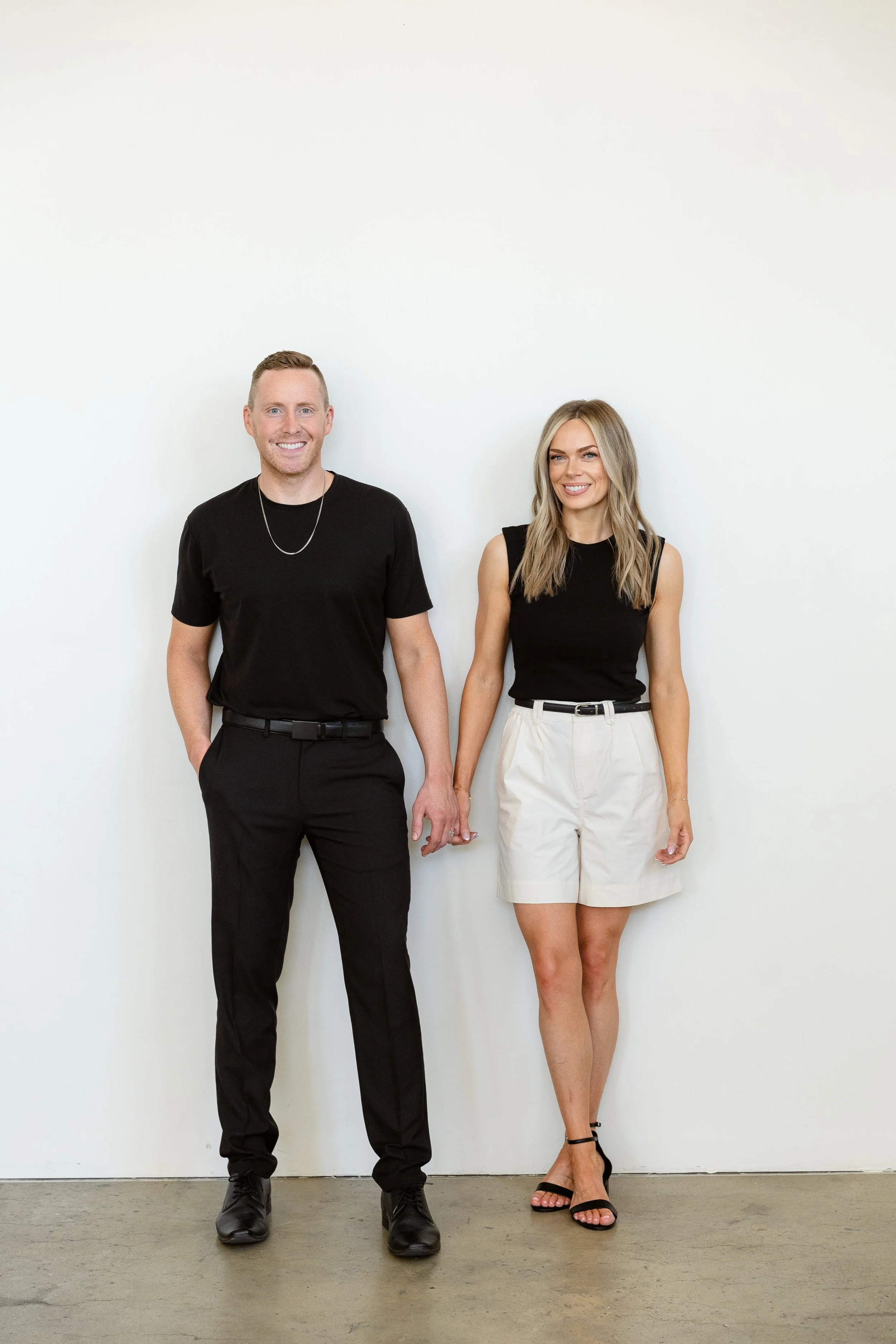 A man and woman holding hands, standing against a plain white wall, dressed in black and white clothes. The man is wearing a black t-shirt and black pants, and the woman is wearing a black sleeveless top and white shorts.