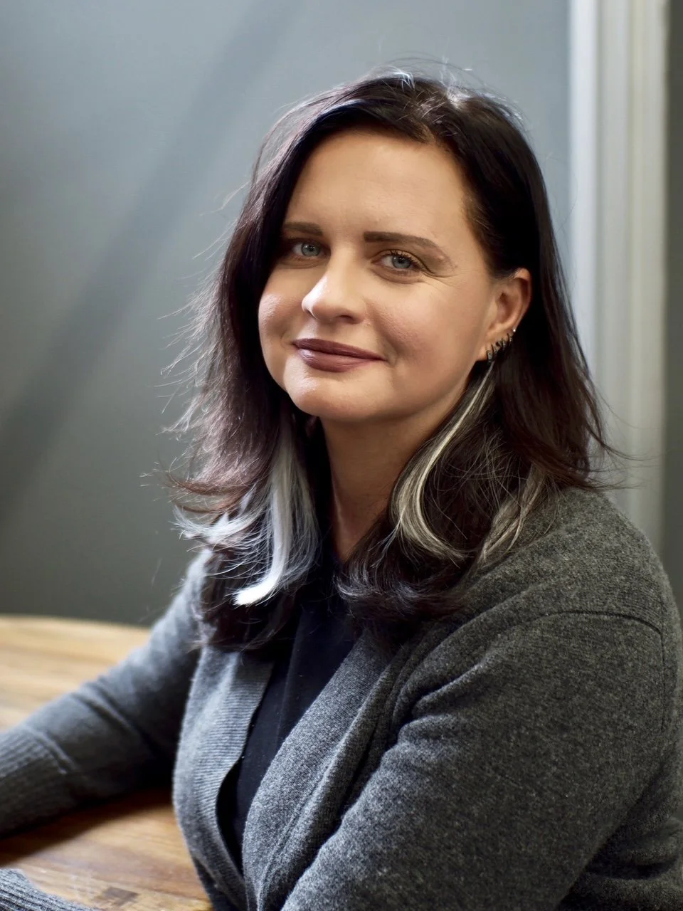 A woman with dark hair and blonde highlights smiling at the camera, sitting at a wooden table in a room with gray walls  This is the social work CE education teacher