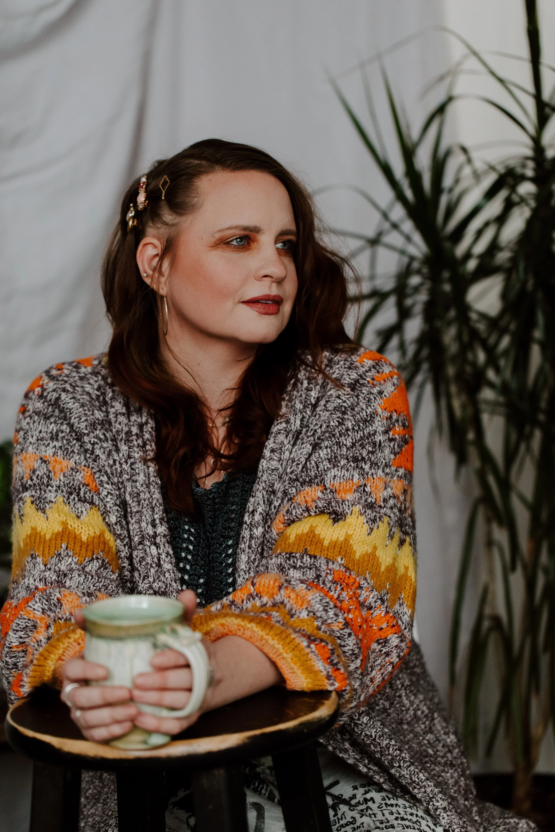 A woman with brown hair and blue eyes sitting at a table, holding a mug, wearing a colorful patterned sweater, with a plant in the background.