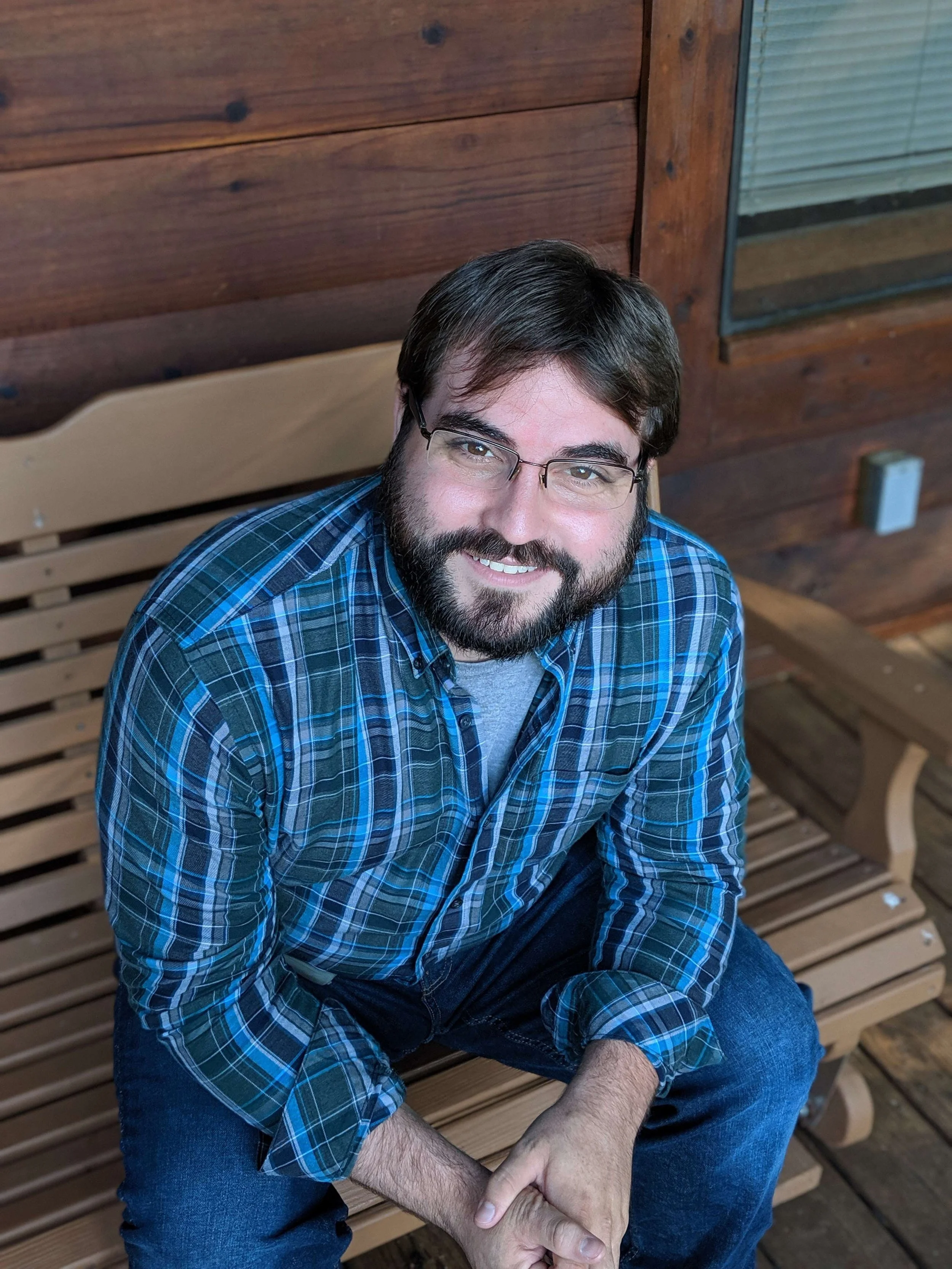 A man with a beard and glasses, wearing a blue plaid shirt, sitting on a wooden bench outdoors, smiling at the camera.