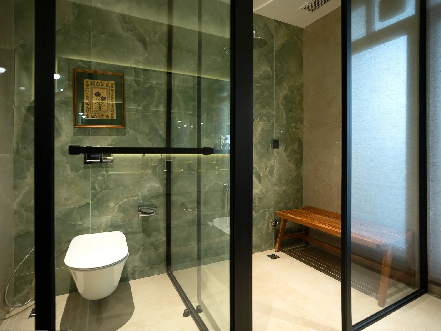 Modern bathroom featuring green marble walls, a white toilet, a wooden bench, and a glass-enclosed shower area, with a framed floral artwork on the wall and frosted window covering.