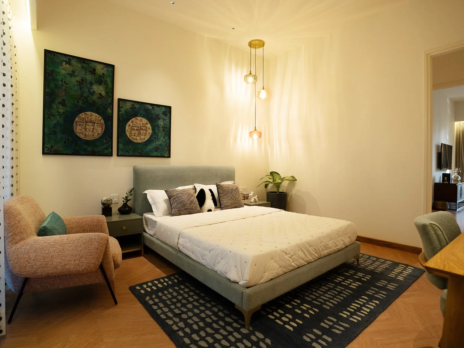 A modern bedroom with a light green bed, two bedside tables, a beige armchair, and a dining table with chairs. Artwork hangs on the wall, and a potted plant is on one of the bedside tables. The room is lit by hanging pendant lights.