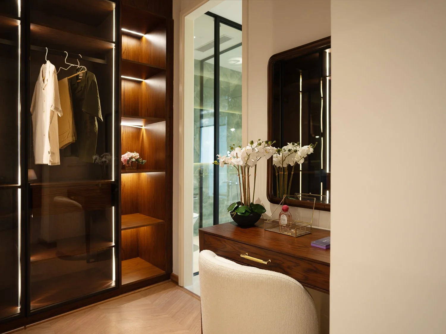 Interior of a bedroom or dressing area with a wooden vanity table, mirror, and decorative items including a potted orchid, perfume, and a book. A wardrobe with glass doors and hanging clothes is visible on the left, and there's a window with a view o