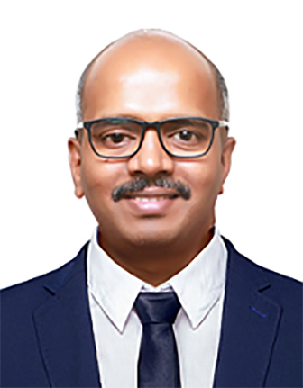 A man with curly dark hair, glasses, and a beard looking slightly to the side. He is wearing a dark green sweater and sitting in a room with a white wall and a plant in the background.