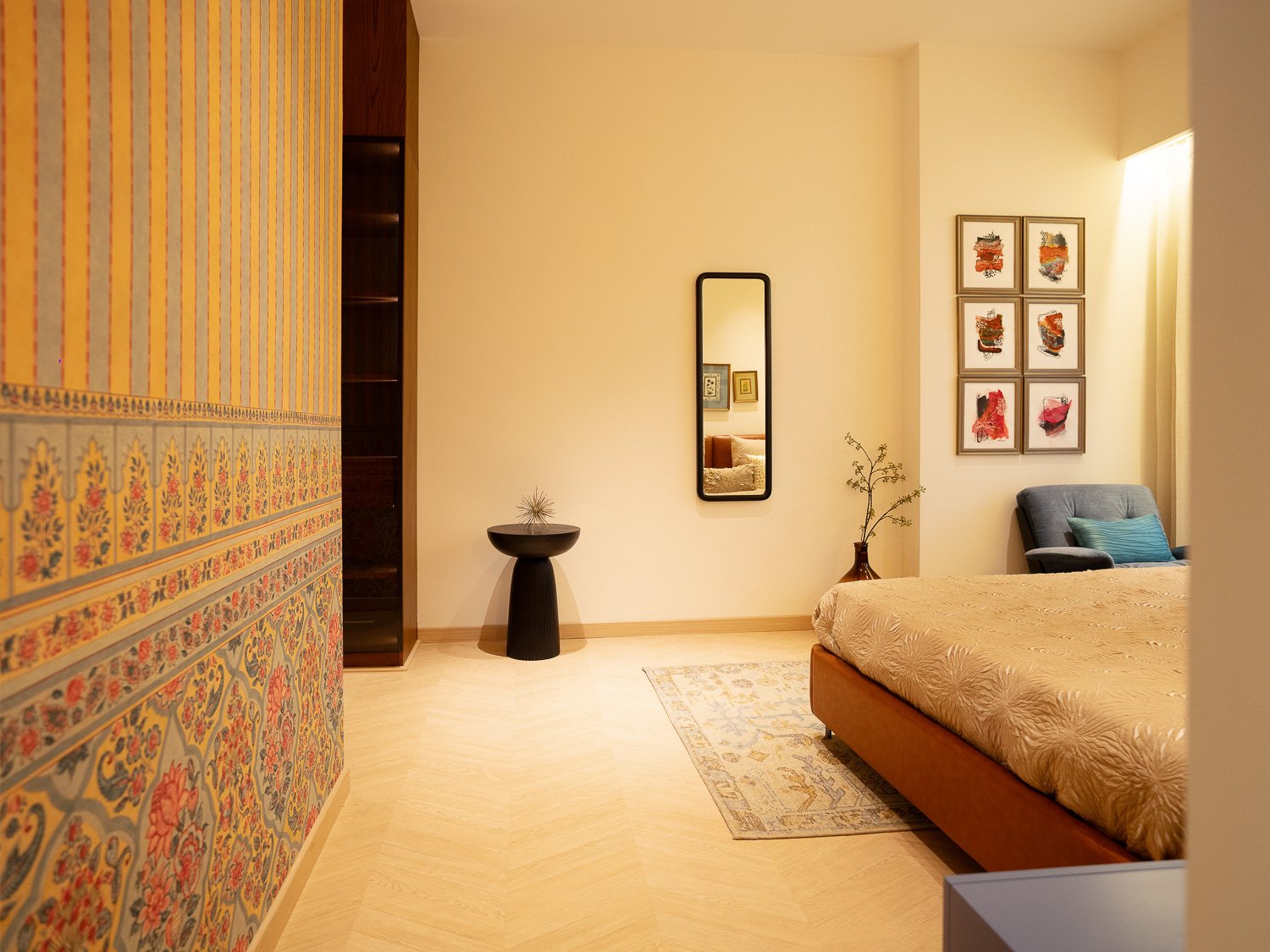 A bedroom with a bed, a mirror on the wall, artwork, a small pillow, and a chair. There's a decorative wall covering on the left and a small table with a plant.